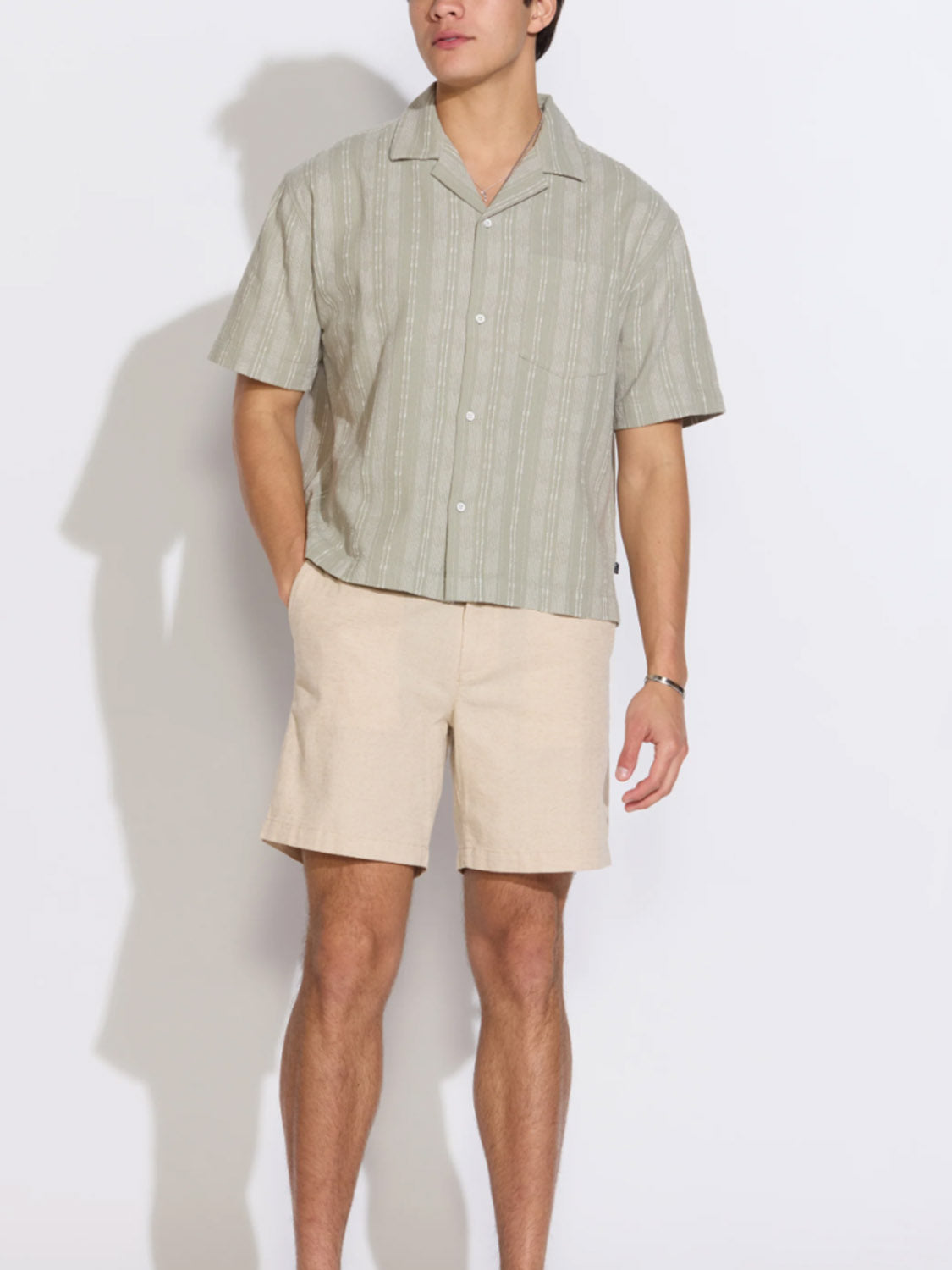 A man stands against a white background wearing beige Civil Society Vito Relaxed Fit Shorts and a light green striped short-sleeve shirt. His left hand is in his pocket, and his face is not visible.