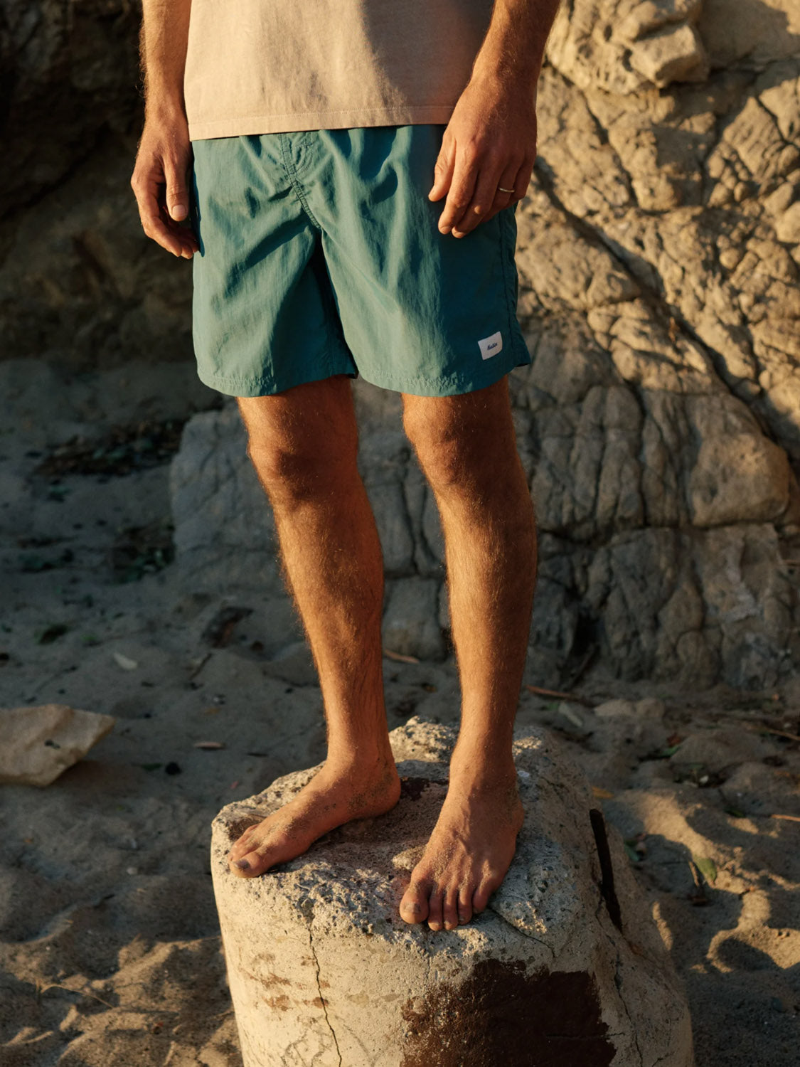 A person wearing a beige shirt and Katin Poolside Volley teal nylon shorts with an elastic waistband stands barefoot on a large rock, their legs and lower torso lit by warm sunlight against a sandy, rocky backdrop.