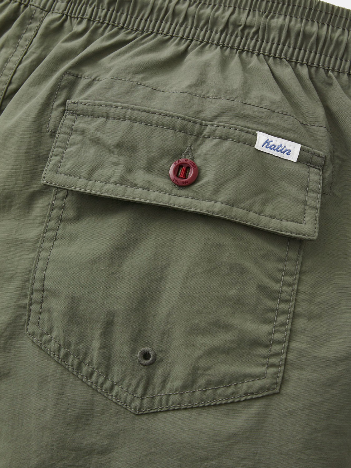 Close-up of the Katin Poolside Volley shorts’ olive green nylon pocket, featuring a red button, small white Katin label above the flap, and elastic waistband—signature details from Katins collection.
