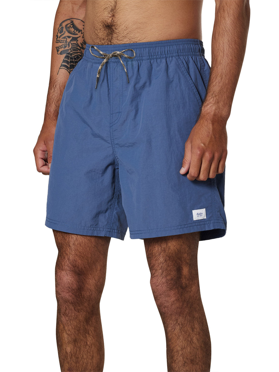 A man wears Katin Poolside Volley swim shorts from Katin in blue, featuring an elastic waistband, side pockets, mesh brief lining, and a small white label on the left leg. His midsection and left forearm tattoo are visible.