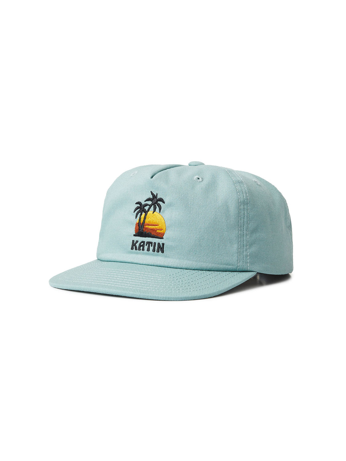 The Katin Voyage Hat is a light blue cotton twill baseball cap with a flat brim, featuring custom embroidery of a yellow sunset and palm trees above KATIN headwear.