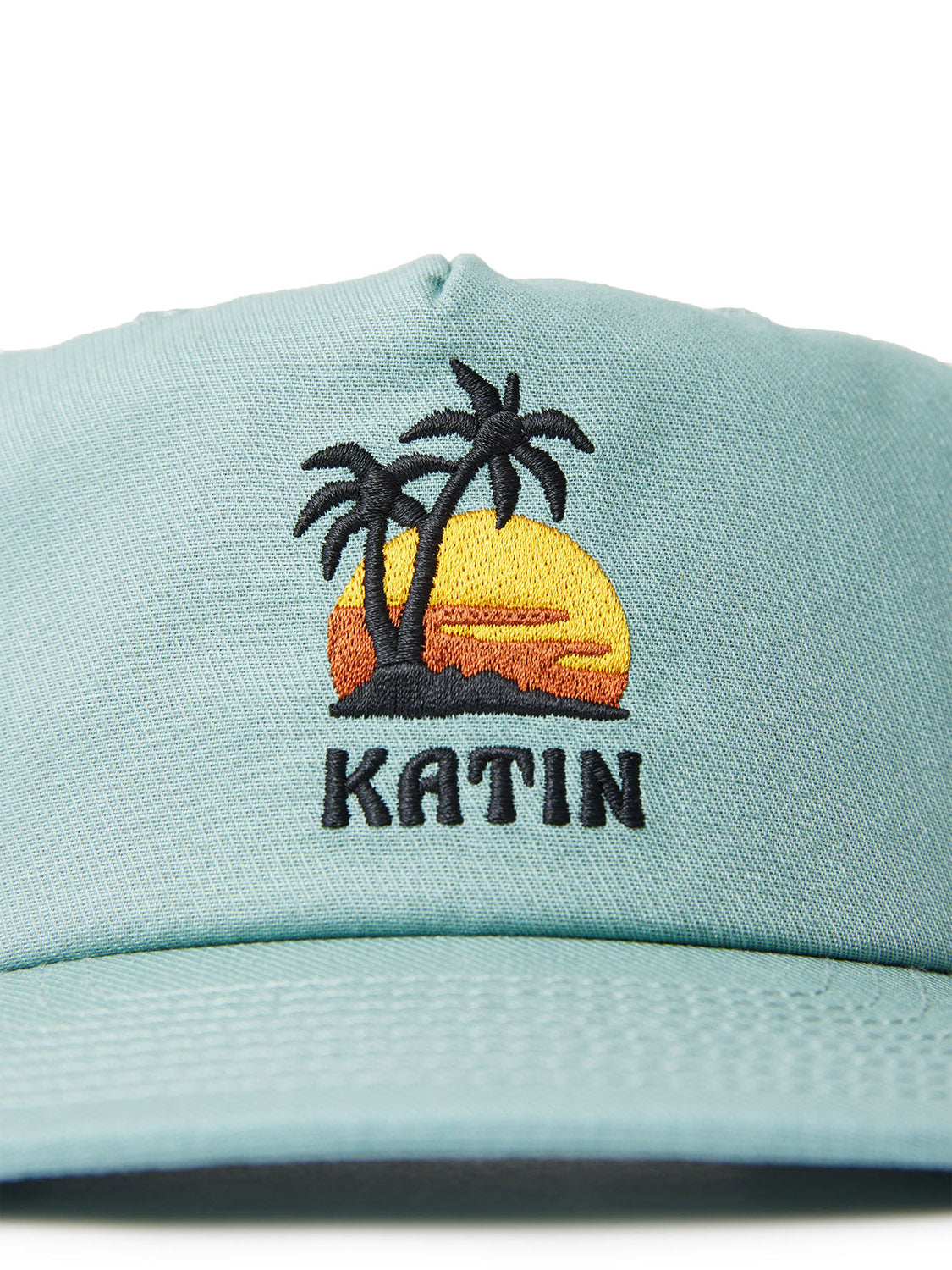 The Katin Voyage Hat, made from cotton twill in light blue, showcases custom embroidery of a sunset and two palm trees with KATIN prominently displayed below.