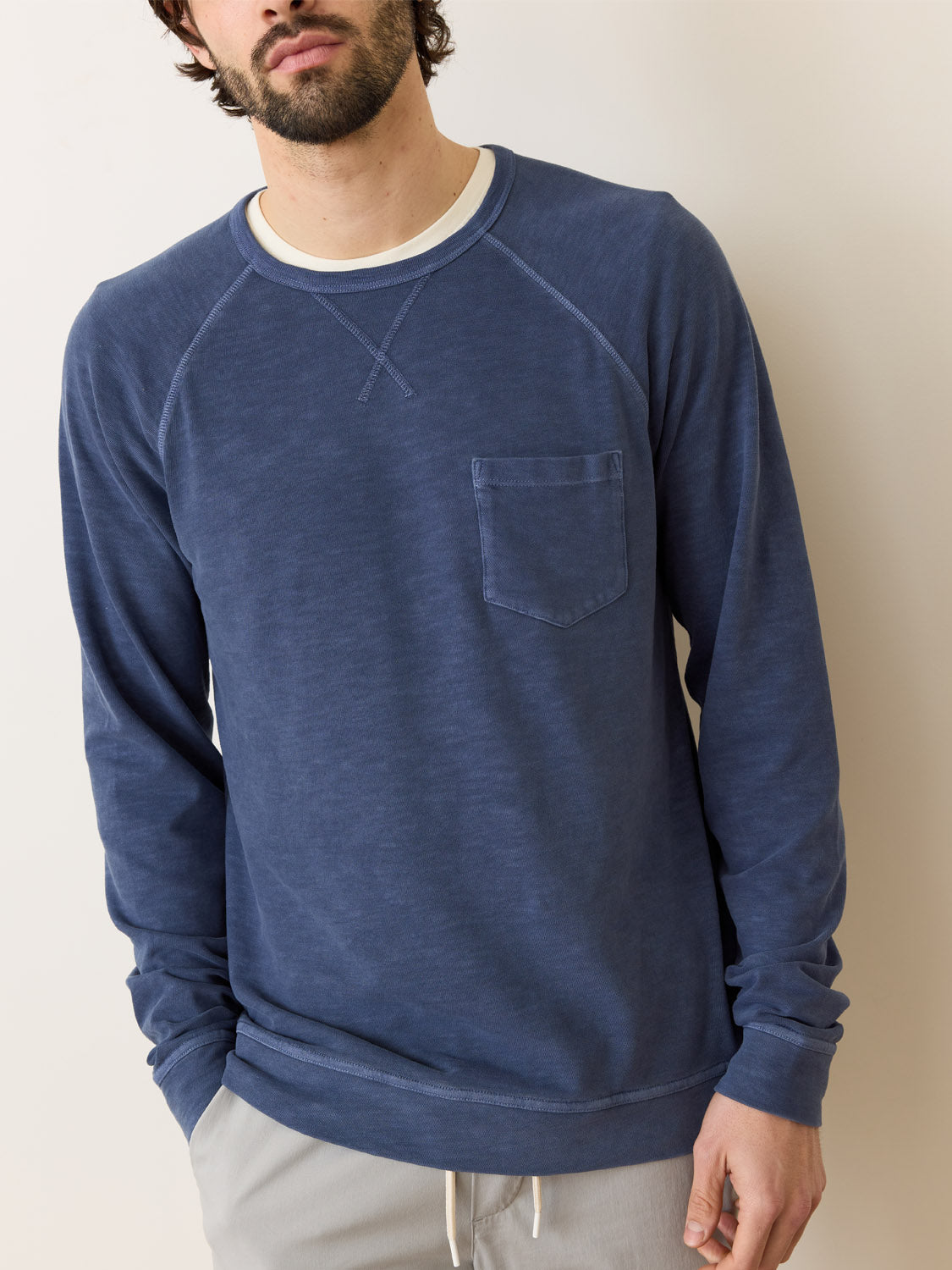 A man stands casually in light gray trousers with hands in his pockets, wearing a blue Marine Layer Vintage Heavy Slub Raglan sweatshirt over a white shirt, its heavyweight material contrasting against the neutral light-colored wall behind him.