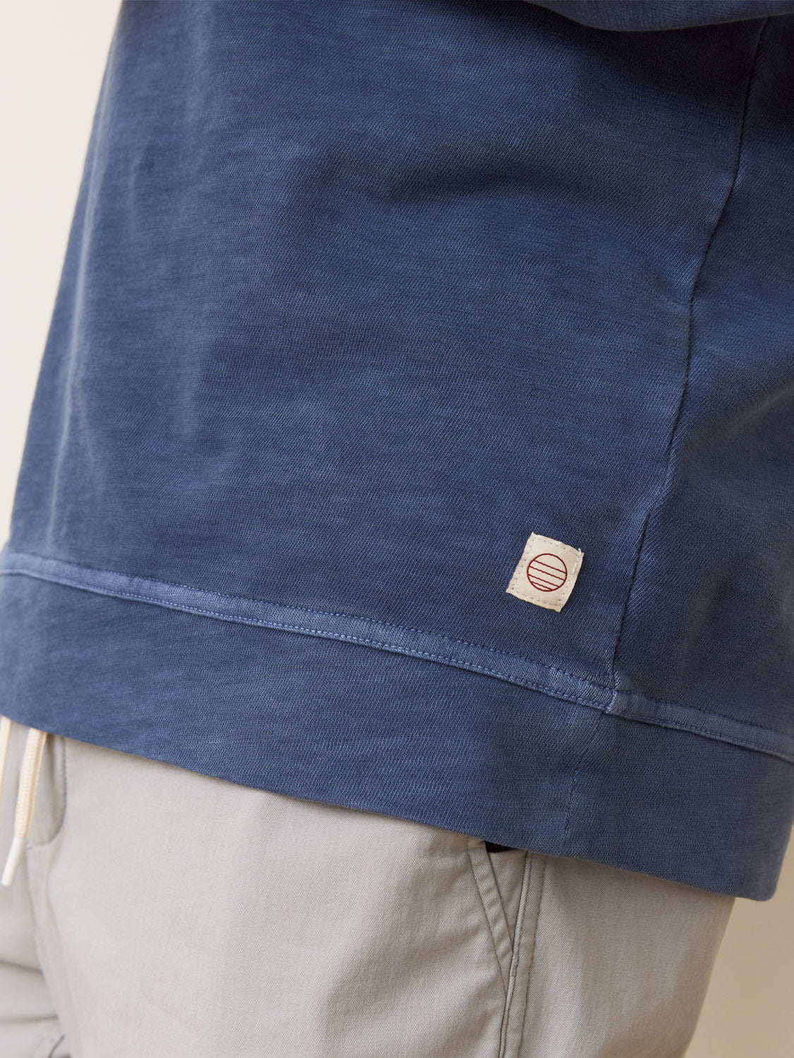 A close-up shows a person in Marine Layers Vintage Heavy Slub Raglan, with its snug fit and small logo tag on the hem, paired with light gray pants. The focus is on the lower part of this blue sweatshirt and pants.