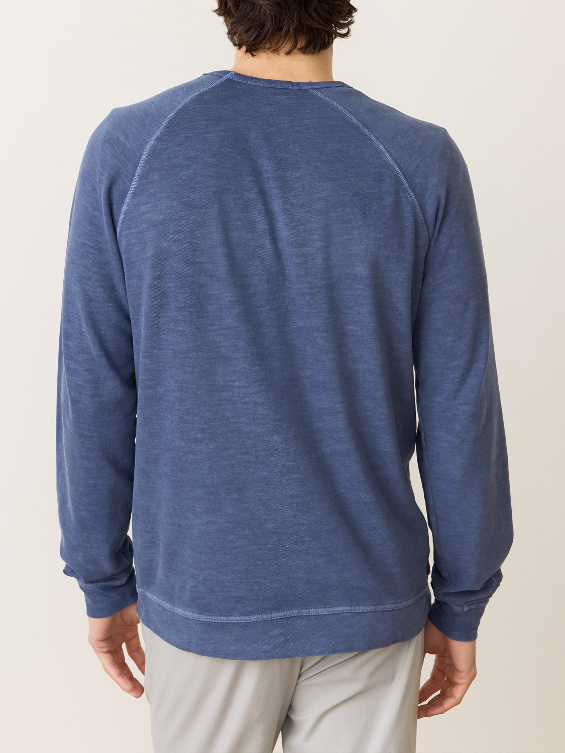 The rear view shows a person wearing the Marine Layer Vintage Heavy Slub Raglan, a long-sleeved faded blue top with raglan sleeves, paired with light gray pants against a plain cream background.