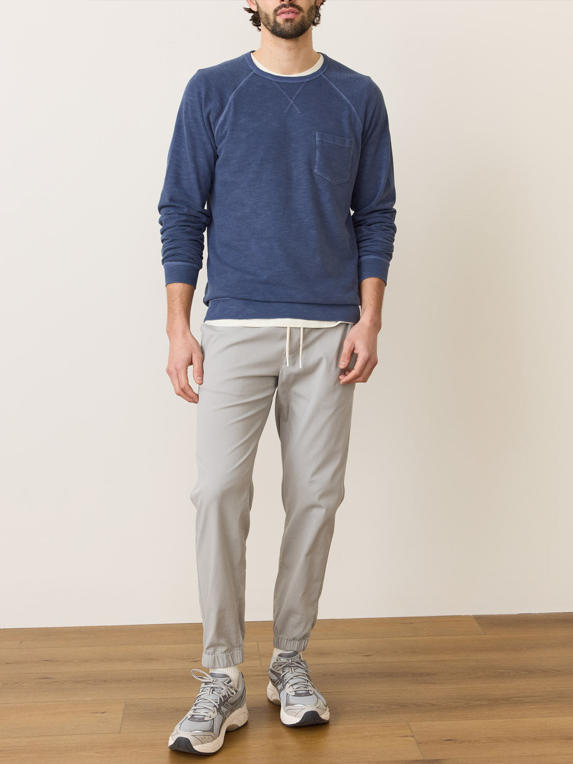 A person stands on a wooden floor against an off-white backdrop, wearing Marine Layer Vintage Heavy Slub Raglan in blue and light gray drawstring pants, along with sneakers. The heavyweight material enhances its appeal, while the persons face remains unseen.
