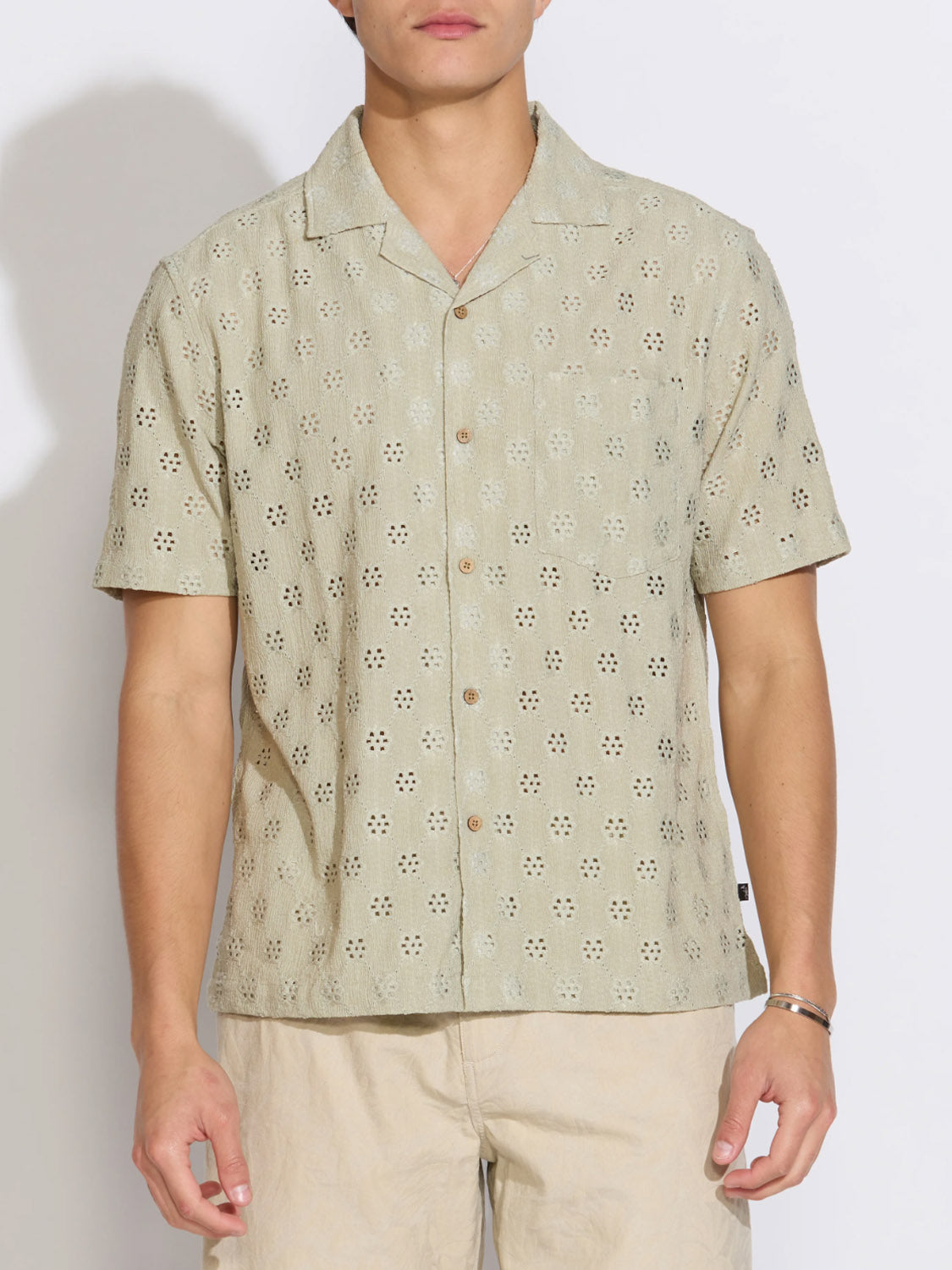 A person wears the Civil Society Wake Eyelet Knit Resort Shirt, a light beige shirt with a subtle daisy eyelet pattern, paired with matching shorts, standing against a plain white background.