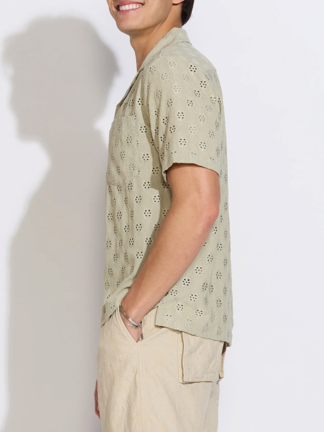 A person stands sideways, smiling, wearing the Civil Society Wake Eyelet Knit Resort Shirt with light beige shorts against a plain white background.