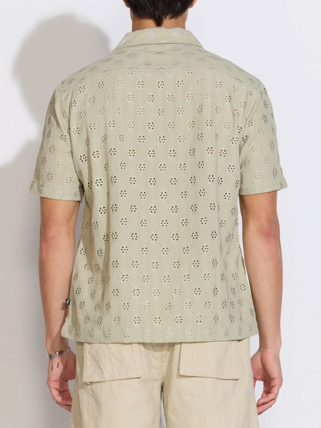 Against a plain white background, a person is seen from the back wearing Civil Society's Wake Eyelet Knit Resort Shirt in light olive green with a daisy eyelet pattern and beige shorts.