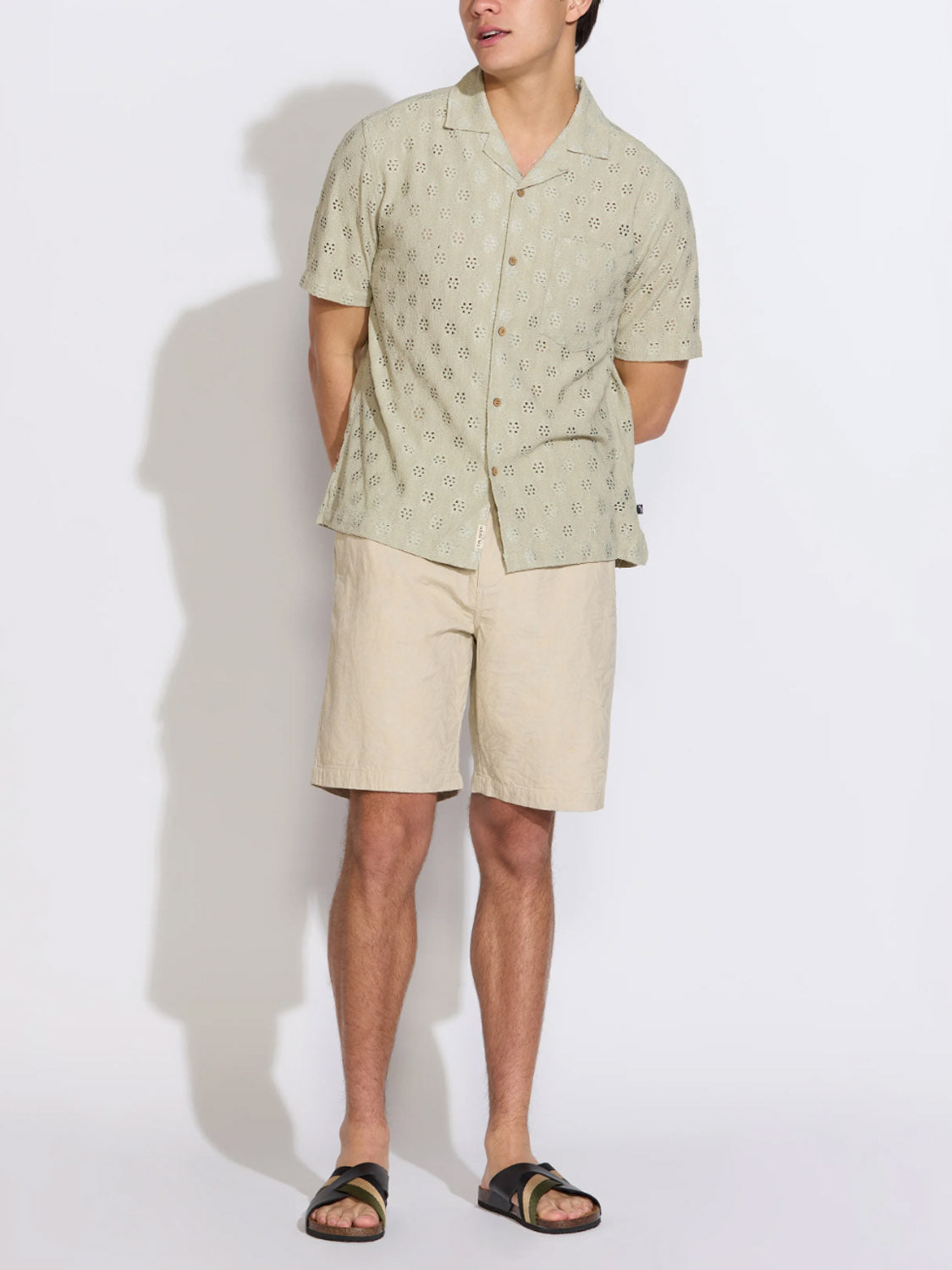 Against a plain background, a person wears the Civil Society Wake Eyelet Knit Resort Shirt with beige shorts and black sandals. Their hands are behind their back and their face is partly out of frame.