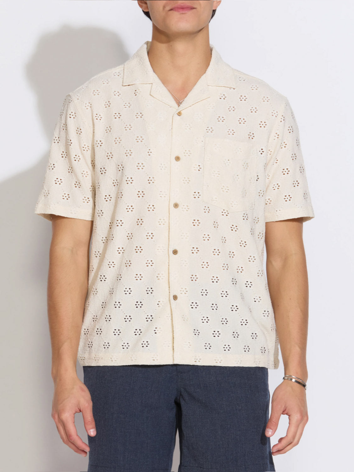 A person wearing the Civil Society Wake Eyelet Knit Resort Shirt, featuring cream-colored short sleeves, daisy eyelet pattern, and chest pocket, with dark pants stands against a plain white background.