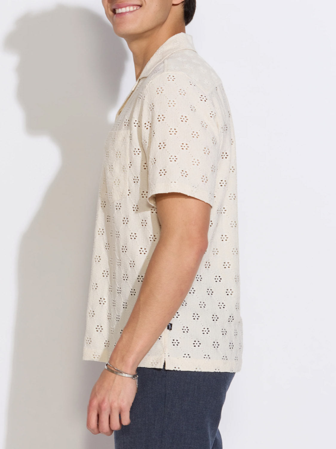 A person in profile smiles while wearing Civil Society’s Wake Eyelet Knit Resort Shirt—short-sleeved and cream-colored—paired with dark pants, standing against a plain white background.