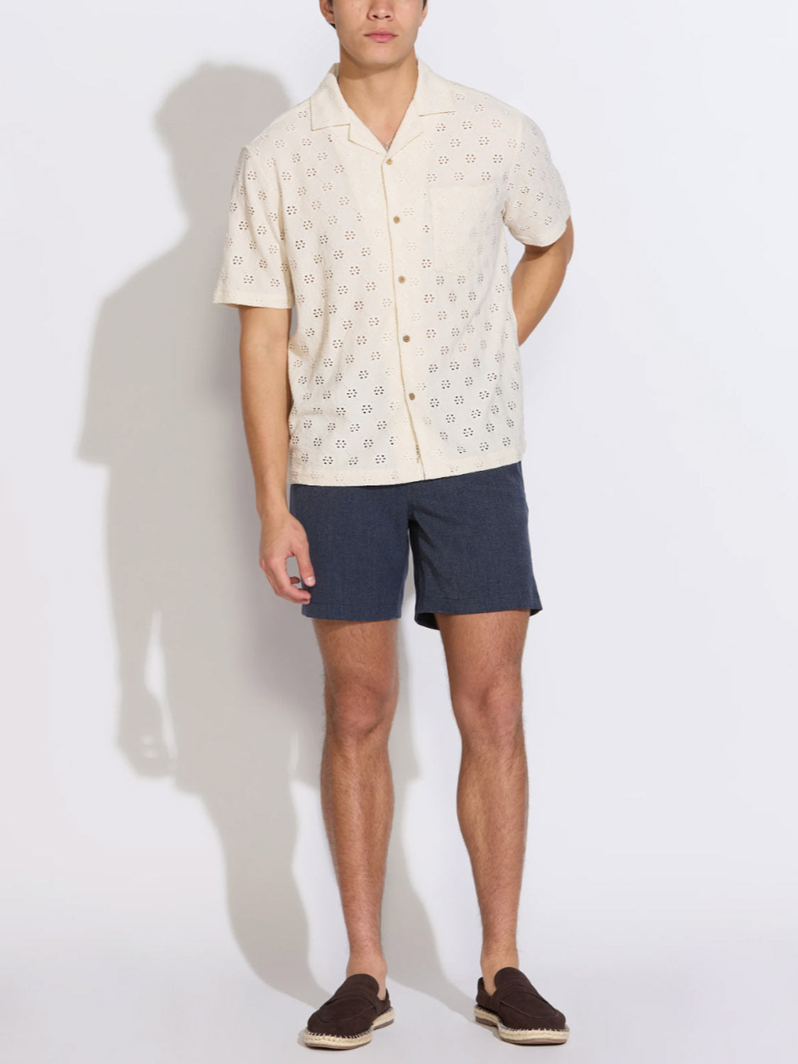 A man stands against a white background wearing the Civil Society Wake Eyelet Knit Resort Shirt, navy shorts, and dark slip-on shoes. One hand is in his pocket and his legs are slightly apart.
