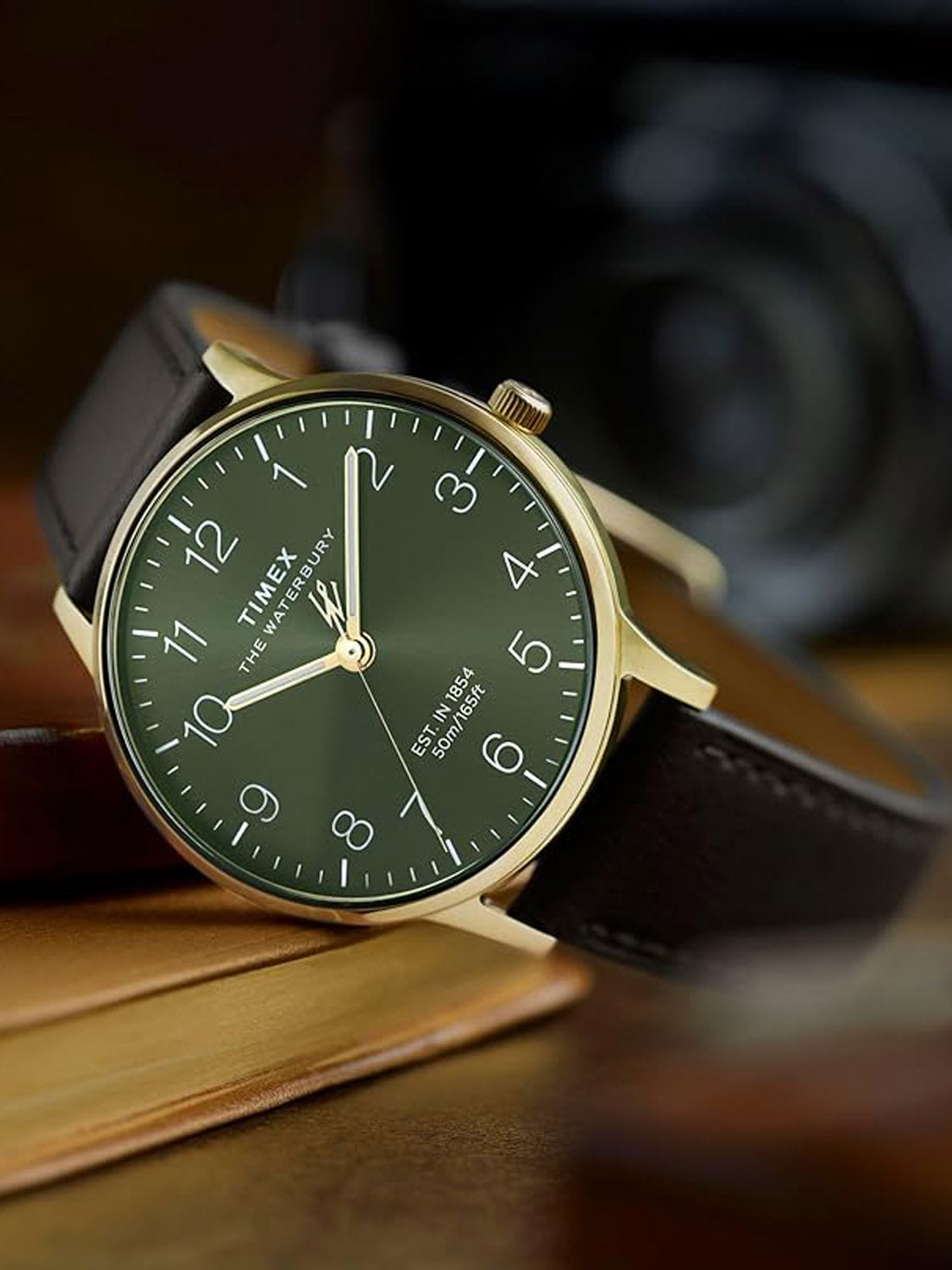 The Timex Waterbury Classic 40mm features a gold-tone stainless steel case, green sunray dial with white numerals, and a black leather strap, shown resting on closed books with a blurred camera in the background.