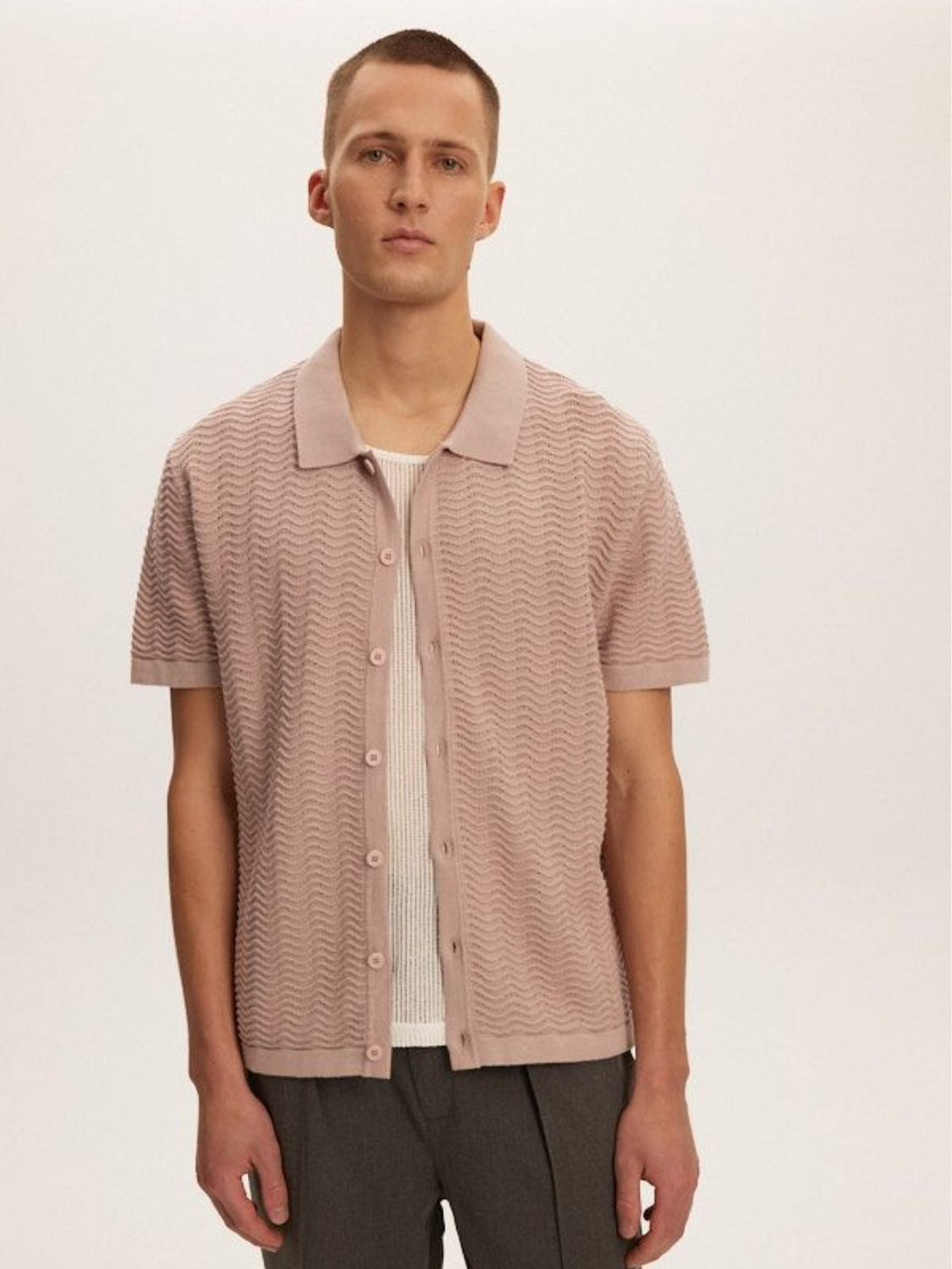 A person stands against a plain background wearing the Kuwalla Wave Knit Polo by Kuwalla, a 100% cotton, short-sleeve button-up shirt in light pink over a white undershirt and gray pants. They have short hair and a neutral expression.