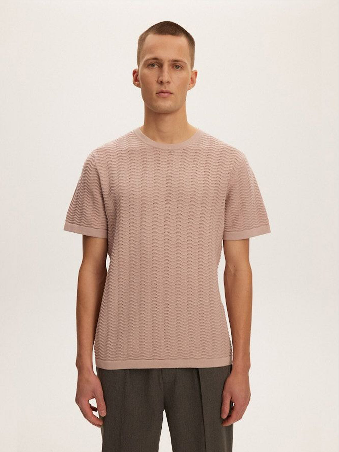A person with short hair is wearing the Kuwalla Wave Knit Tee in light pink paired with gray pants. This stylish outfit from Kuwalla is lightweight, breathable, and features a textured wave pattern on a plain, light gray background.