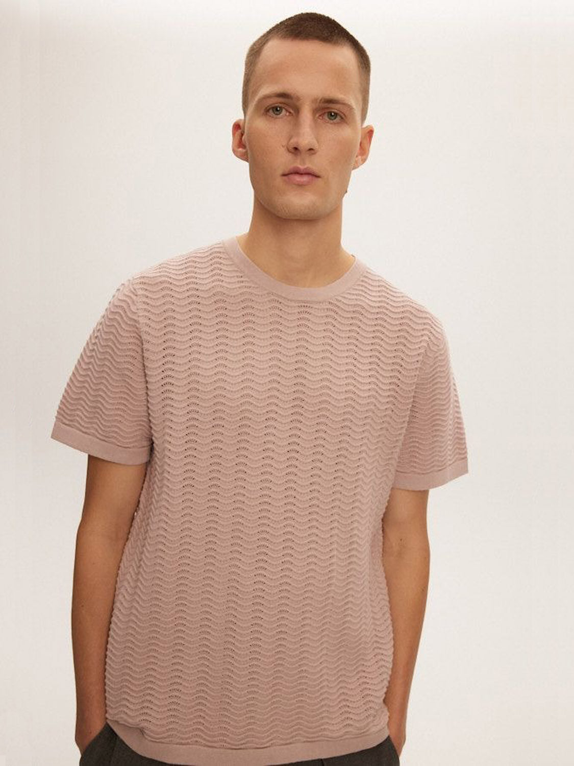 A person with short hair wears a Kuwalla Wave Knit Tee in light pink, featuring a lightweight and breathable texture. They stand against a plain, light-colored background with a neutral expression and hands in pockets.