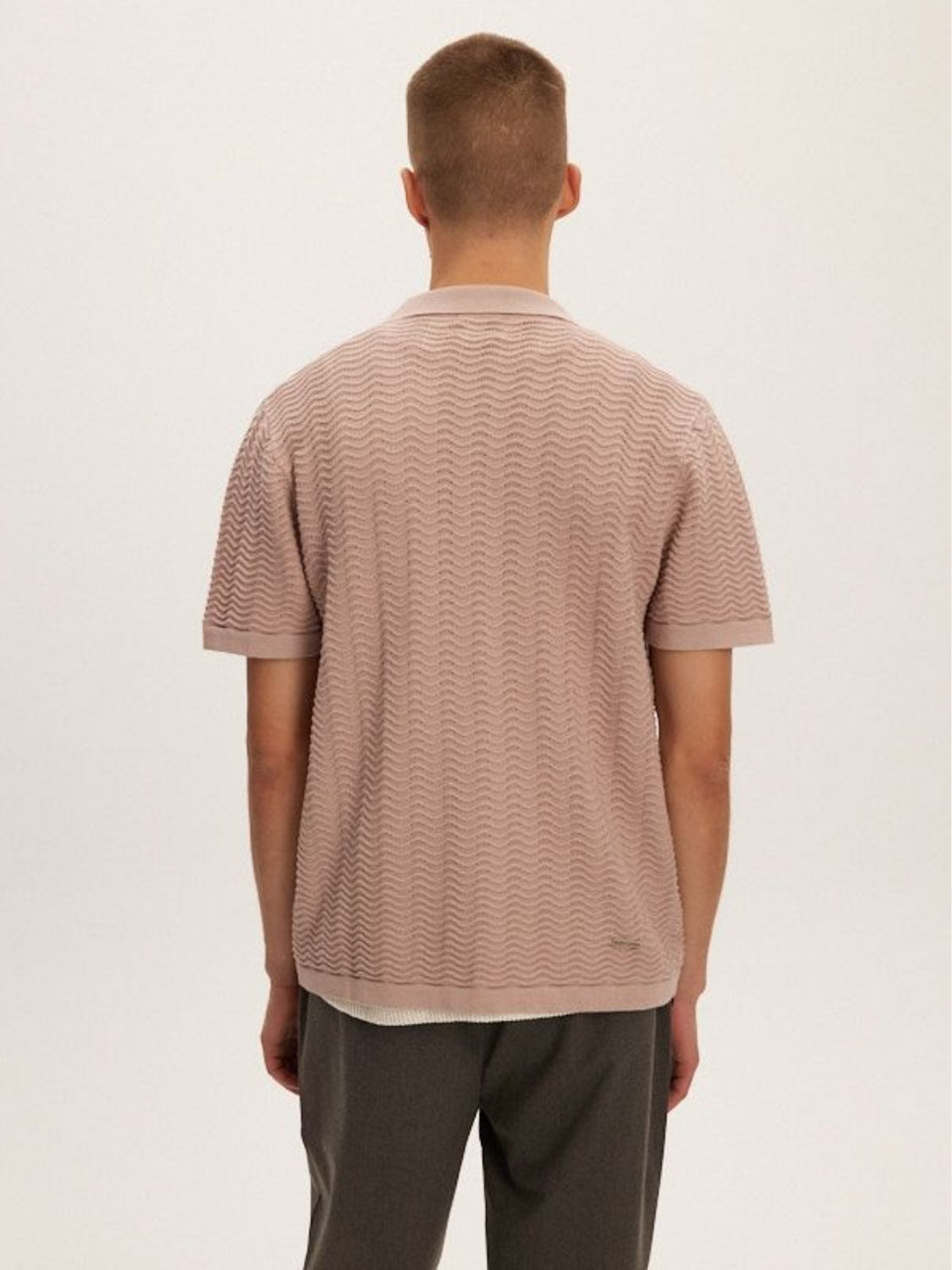 A person with short hair stands with their back to the camera, wearing a Kuwalla Wave Knit Polo and dark pants. The shirt is a button-up, light brown, textured knit made from 100% cotton. The plain background is light-colored.
