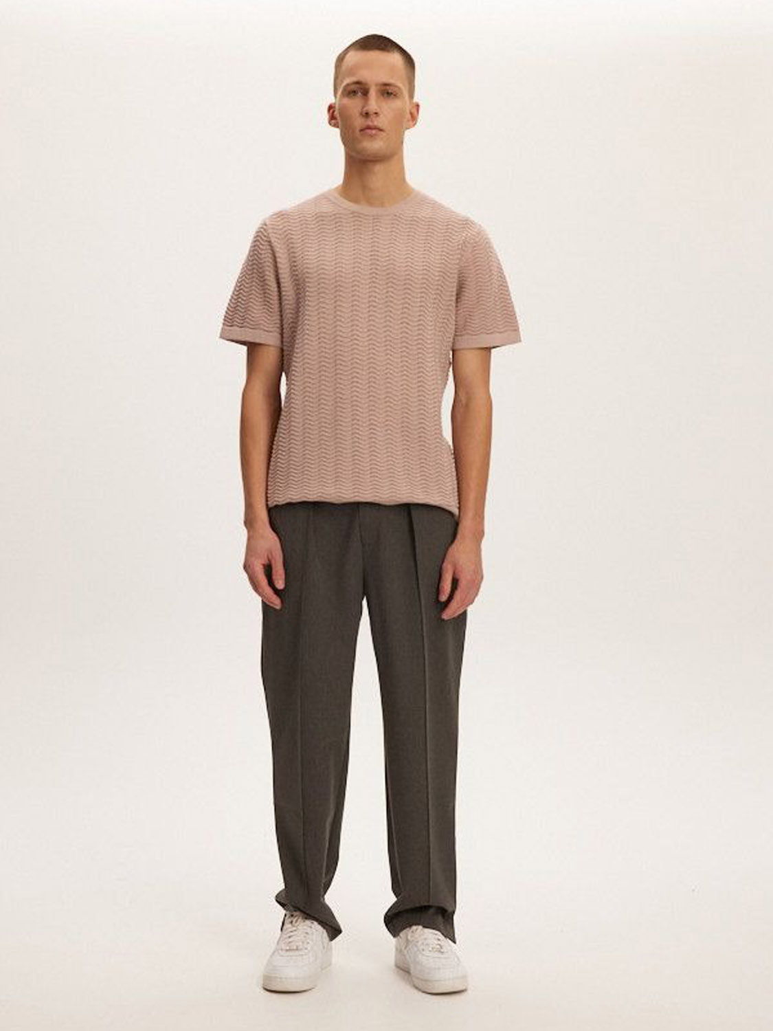A person stands against a plain background wearing a Kuwalla Wave Knit Tee in pink, paired with dark gray pants and white sneakers. The lightweight, breathable outfit matches their neutral expression as they gaze ahead.