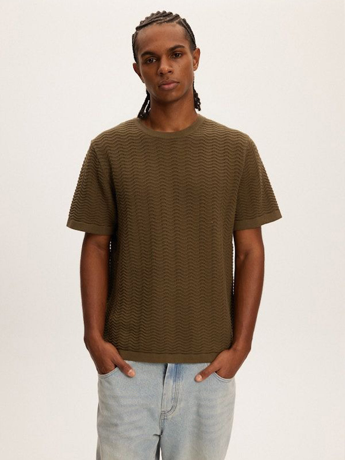 A person with braids and a neutral expression stands against a plain, light background, wearing Kuwallas lightweight and breathable Wave Knit Tee in olive green paired with light blue jeans.