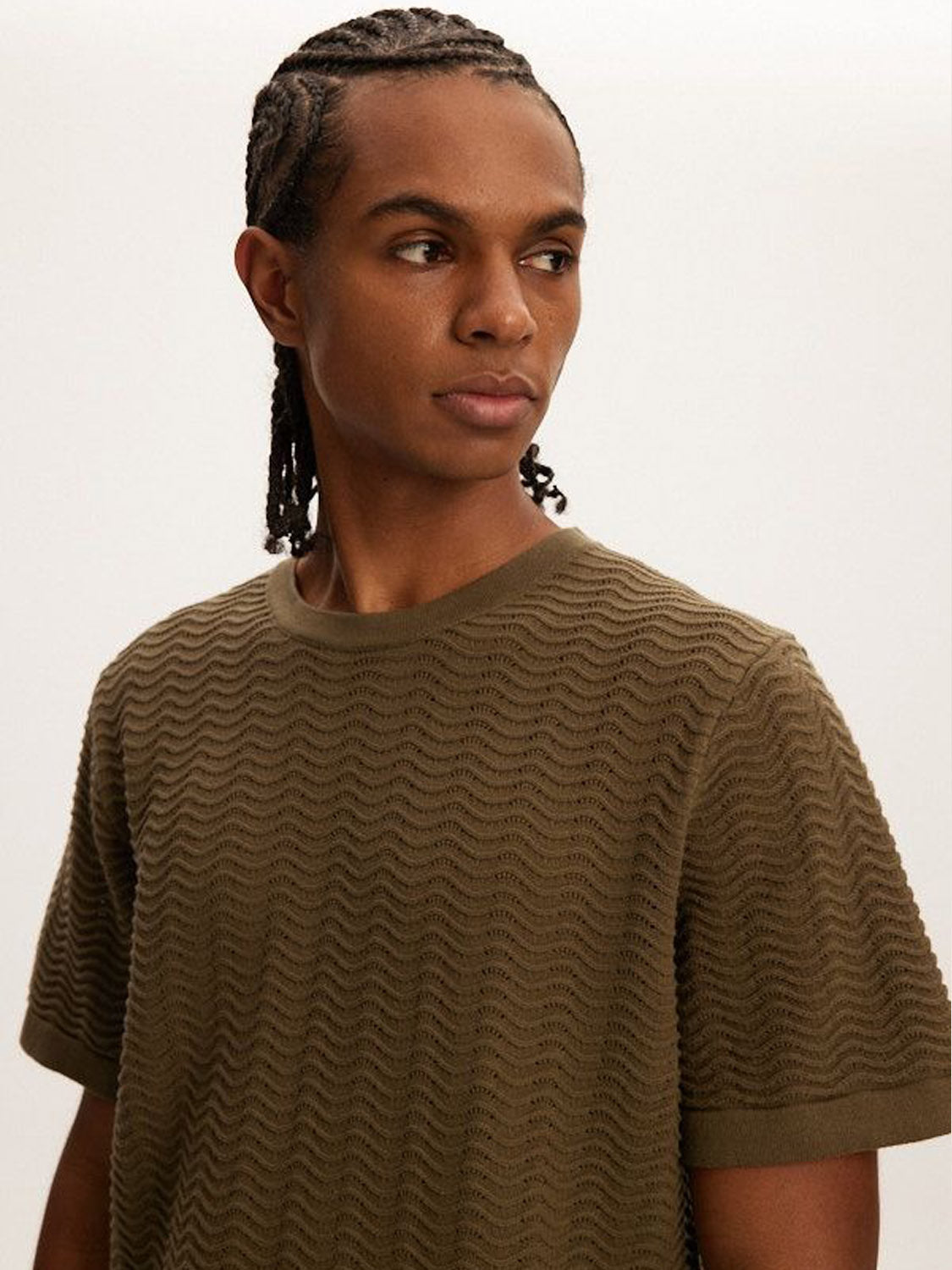 A person with braided hair sports the Kuwalla Wave Knit Tee in versatile olive green, glancing sideways against a plain backdrop.