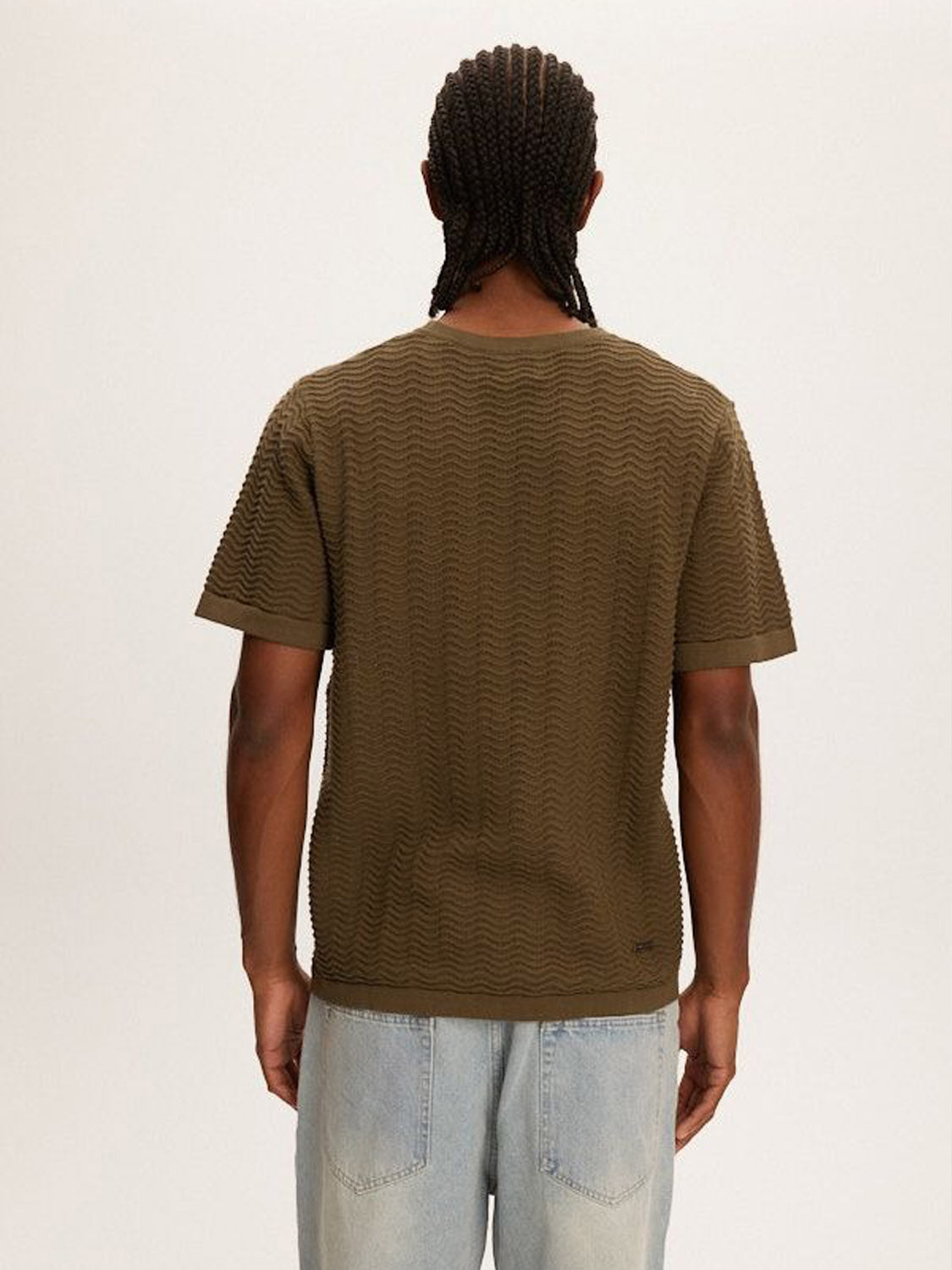A person with braided hair wears the Kuwalla Wave Knit Tee, a versatile, lightweight, breathable olive green t-shirt by Kuwalla, paired with light blue jeans against a plain background.