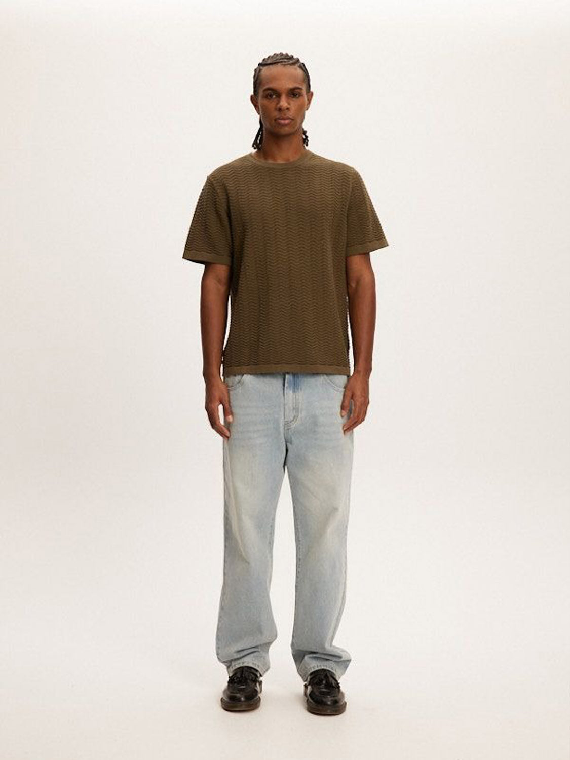 A person with braided hair stands against a plain background wearing the Kuwalla Wave Knit Tee, light blue jeans, and black shoes. The ensemble is versatile, lightweight, and breathable as they face forward with a neutral expression.