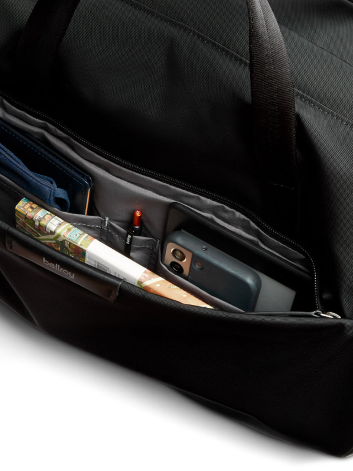 A close-up of the Bellroy Classic Weekender - 45L reveals a minimalist duffel bag with organized inner pockets holding a rolled-up magazine, blue wallet, pen, and smartphone.