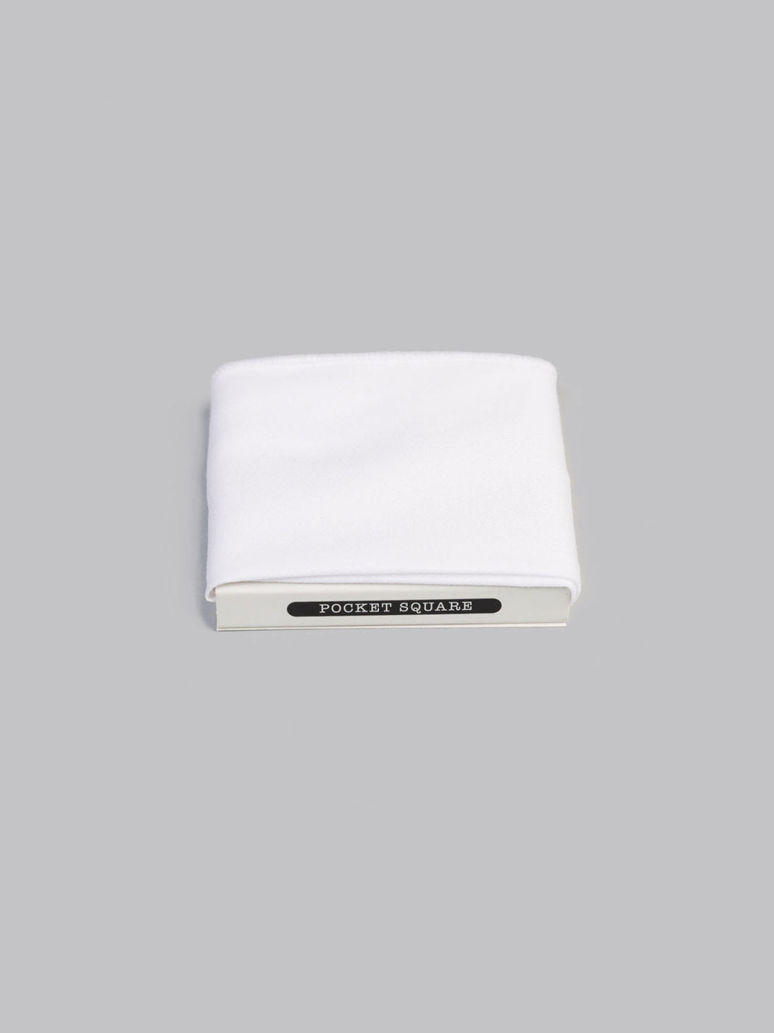 A neatly folded White Cotton Pocket Square by ID Menswear is displayed against a plain light gray background, partially tucked into packaging labeled Pocket Square.