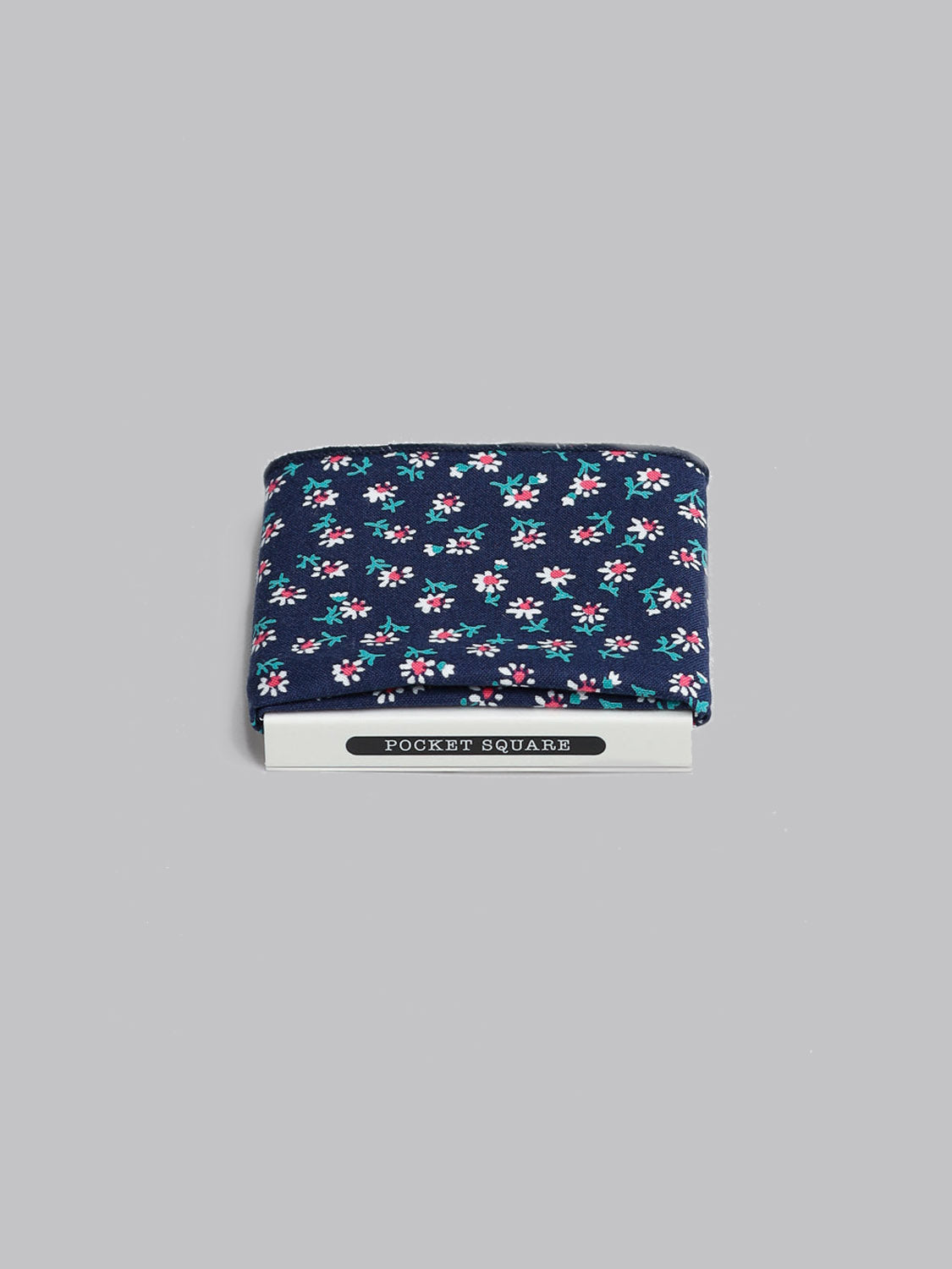The White Bloom Pocket Square by ID Menswear is handmade in Brooklyn from 100% cotton, featuring a navy blue base with small pink, white, and green floral patterns. It’s neatly folded on a branded card against a light gray background.