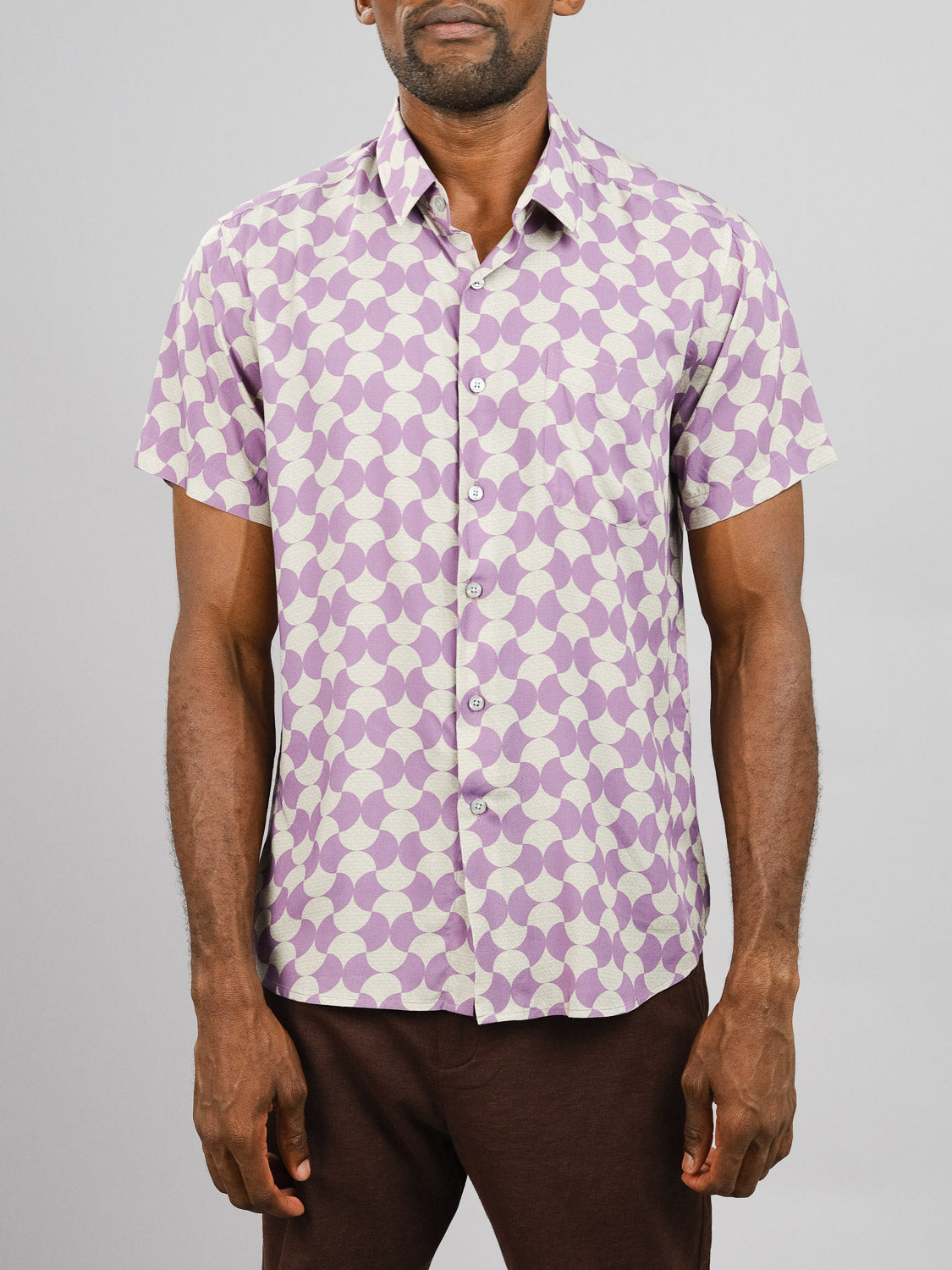 A man wears the Wiggle Printed Rayon Shirt by ID Menswear, featuring a vibrant purple and white geometric design, paired with brown pants, standing against a plain light gray background.