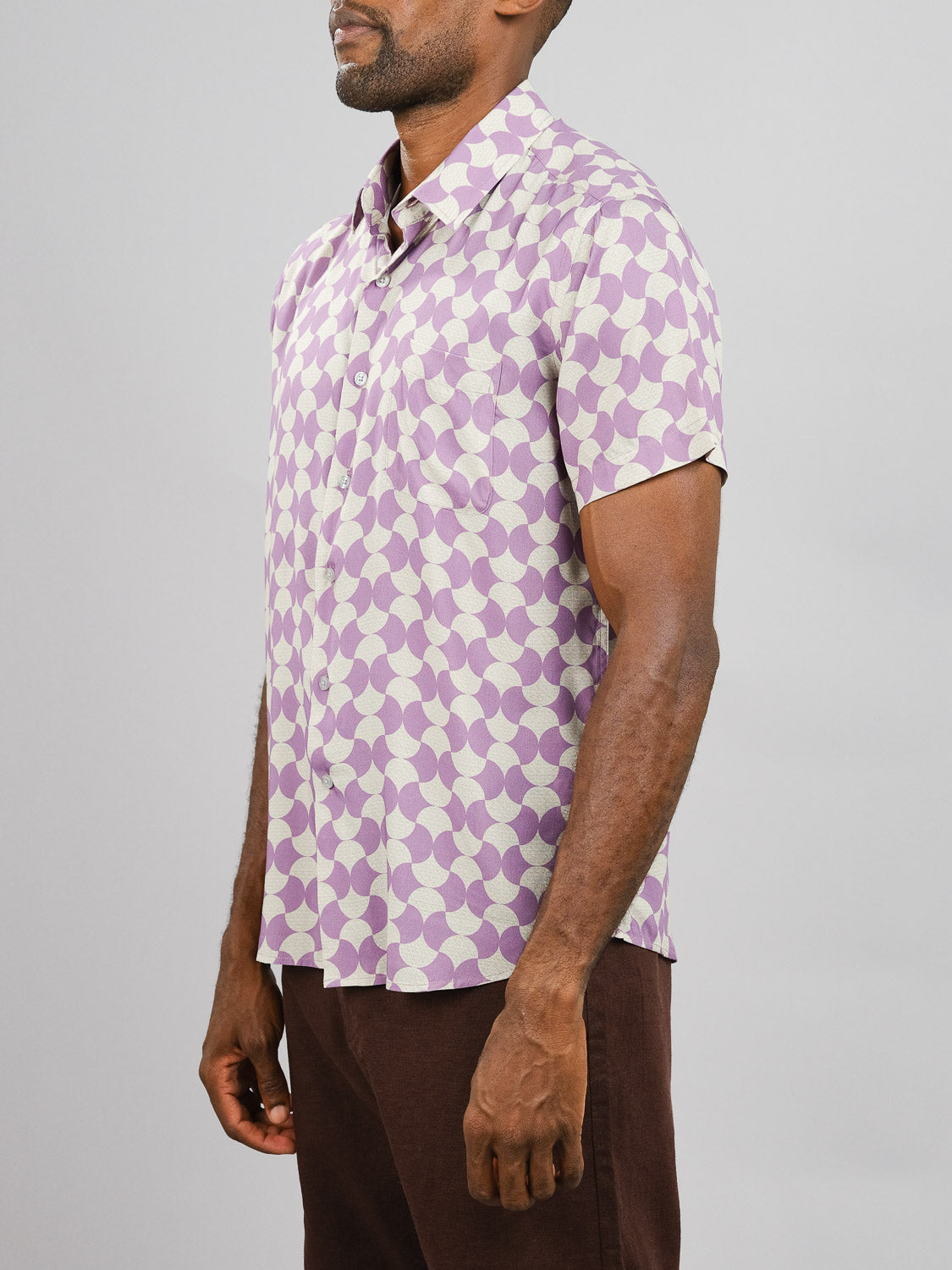 A man wears the Wiggle Printed Rayon Shirt by ID Menswear, a short-sleeve shirt with a light purple and cream geometric pattern, paired with dark brown pants, standing against a plain gray background.