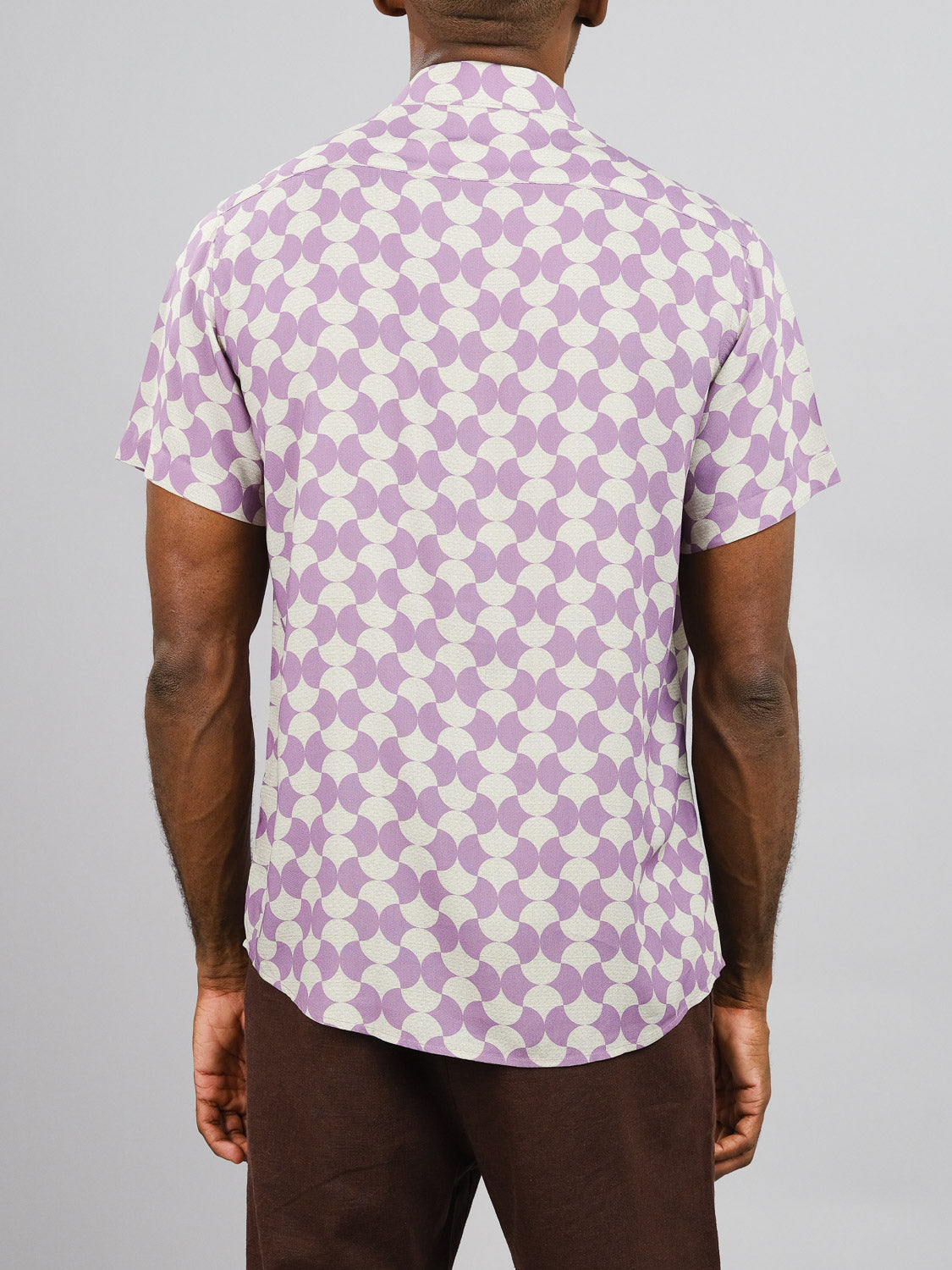 A person stands facing away, wearing the Wiggle Printed Rayon Shirt by ID Menswear, featuring vibrant purple and white geometric prints and short sleeves, paired with brown pants against a plain gray background.