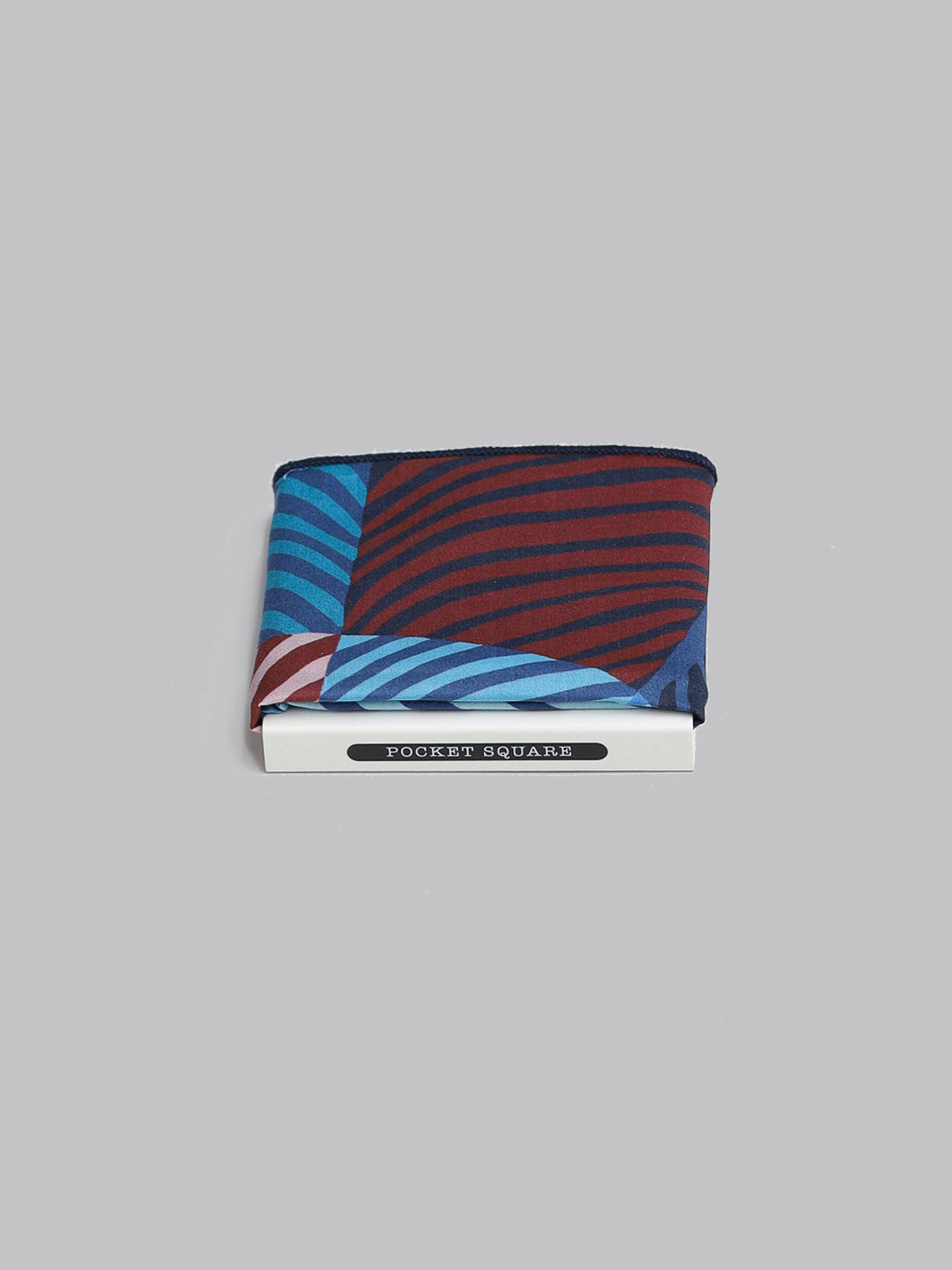 The Liberty™ Wilde Pocket Square by ID Menswear, made from Tana Lawn cotton with blue and red diagonal stripes, is folded atop a small white box labeled POCKET SQUARE on a plain gray background.