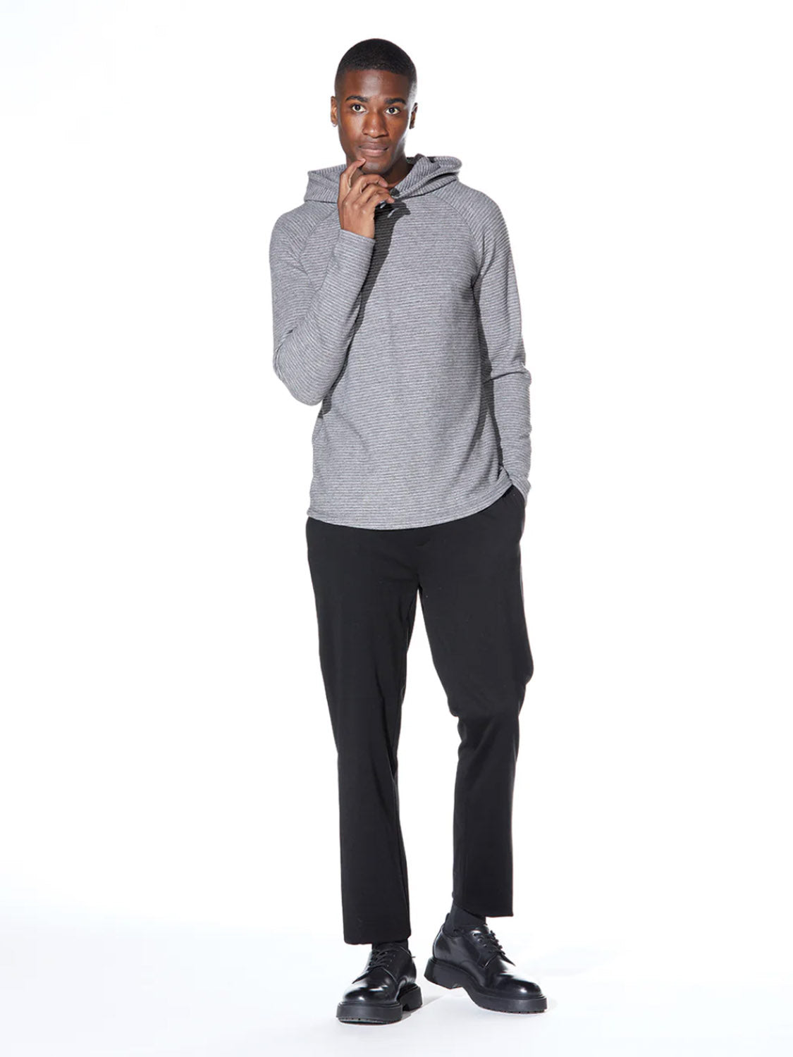 A man poses against a white background wearing the Civil Society Wolfe Waffle Knit Hoodie in gray heather, paired with black pants and black dress shoes. One hand touches his chin while the other rests in his pocket.