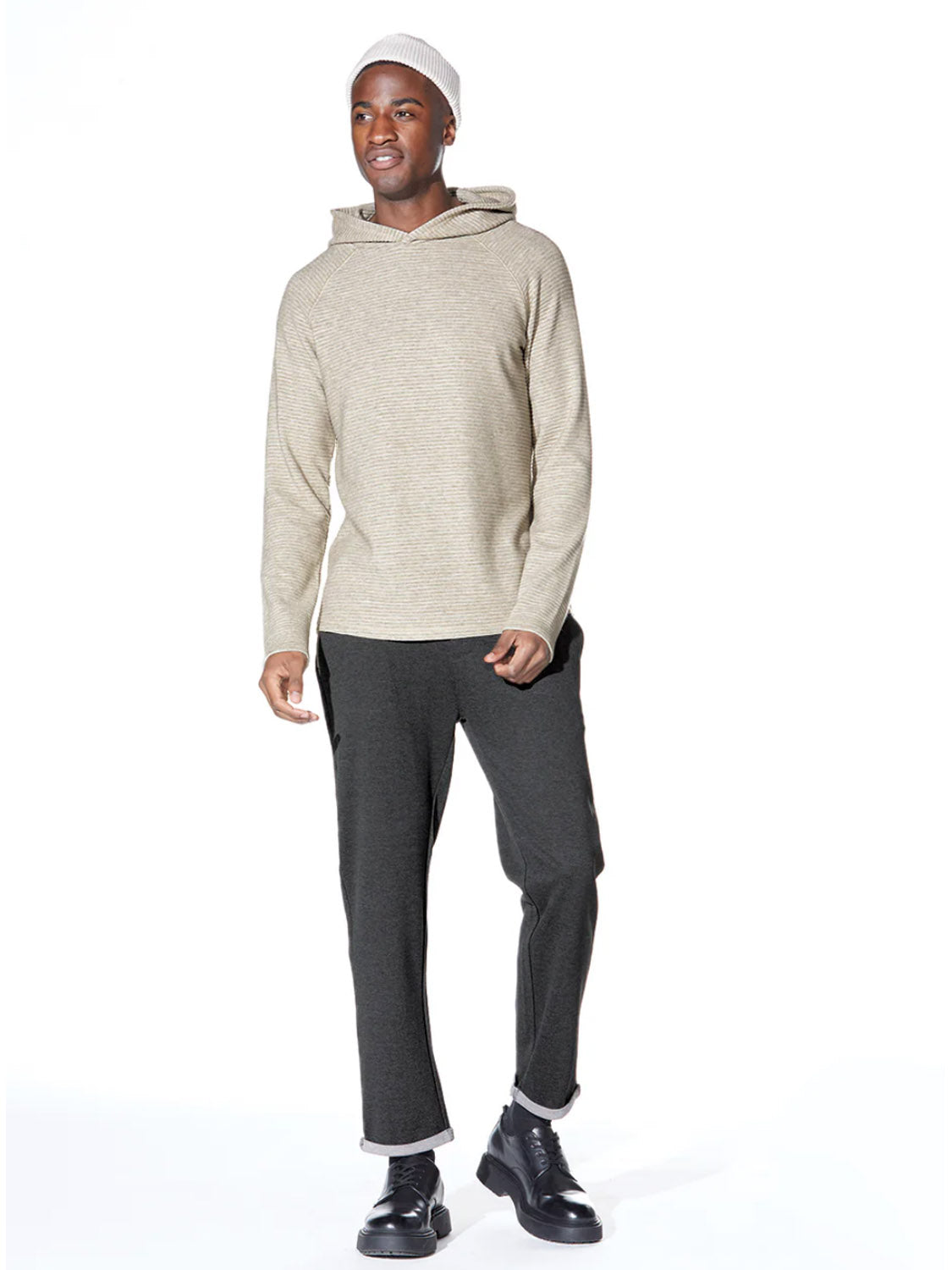 A man wearing a Civil Society Wolfe Waffle Knit Hoodie, dark gray pants, black boots, and a light-colored knit beanie stands against a plain white background.