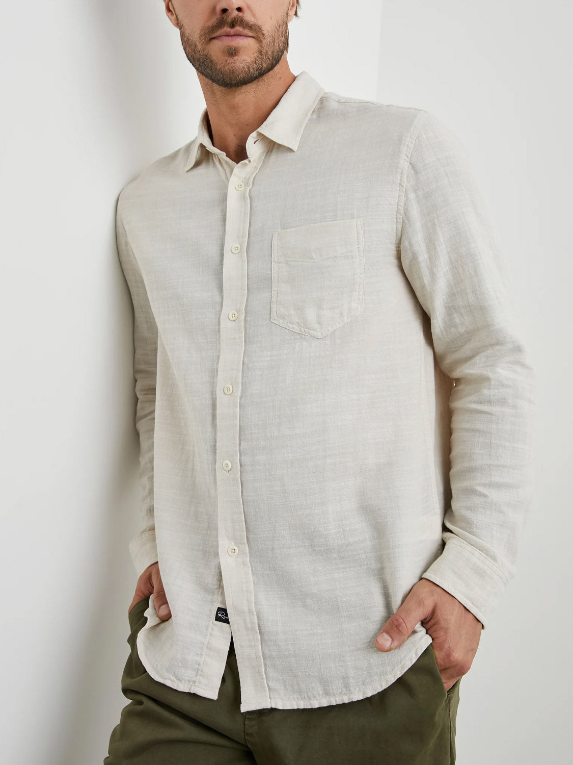 A man wearing a Rails Wyatt Shirt, a classic light beige button-down made from cotton, stands against a white wall. His left hand is in the pocket of his olive green pants as he looks slightly to the side, captured from the waist up.