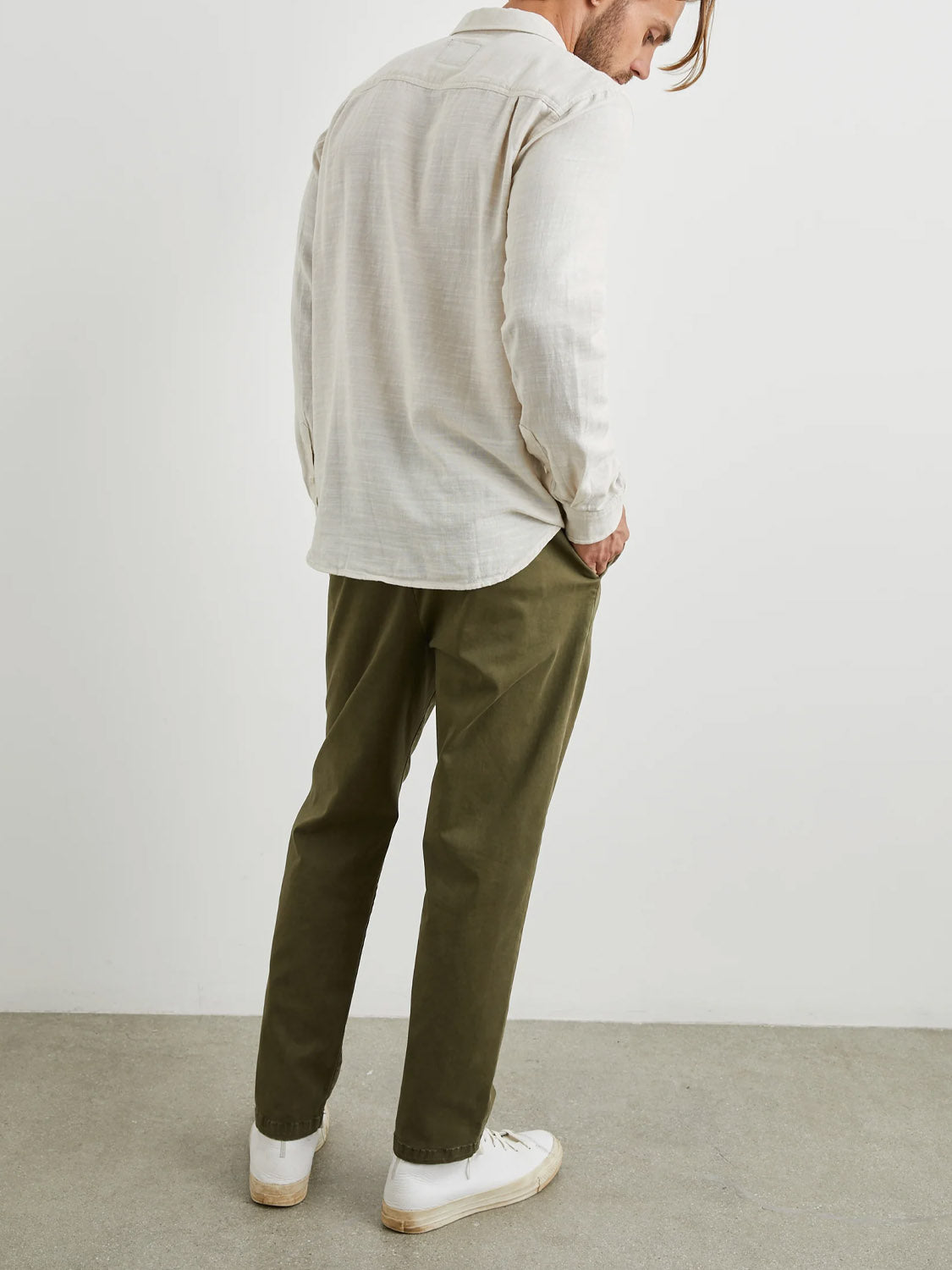 A man stands facing away in a beige Rails Wyatt Shirt, soft cotton fabric, olive green pants, and white sneakers. His left hand is in his pocket. The background shows a plain white wall and gray floor.