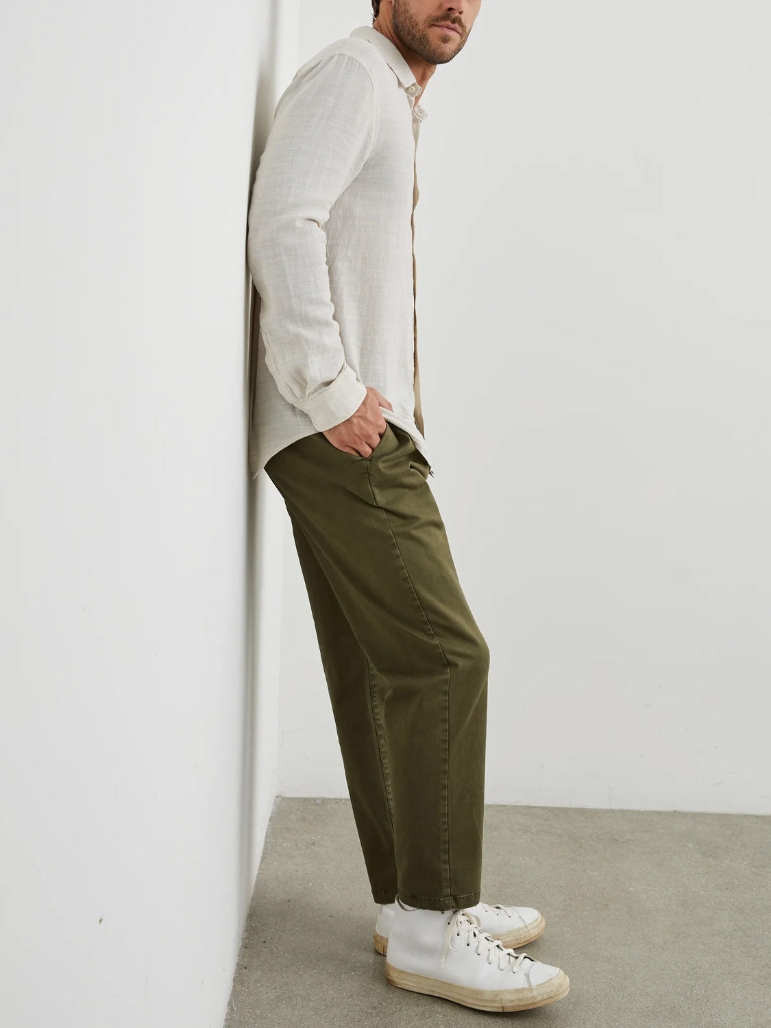Someone leans against a wall in a Rails Wyatt Shirt and olive green pants, paired with white high-top sneakers. The setting is a plain, light-colored room with a concrete floor, exuding an effortlessly relaxed vibe.