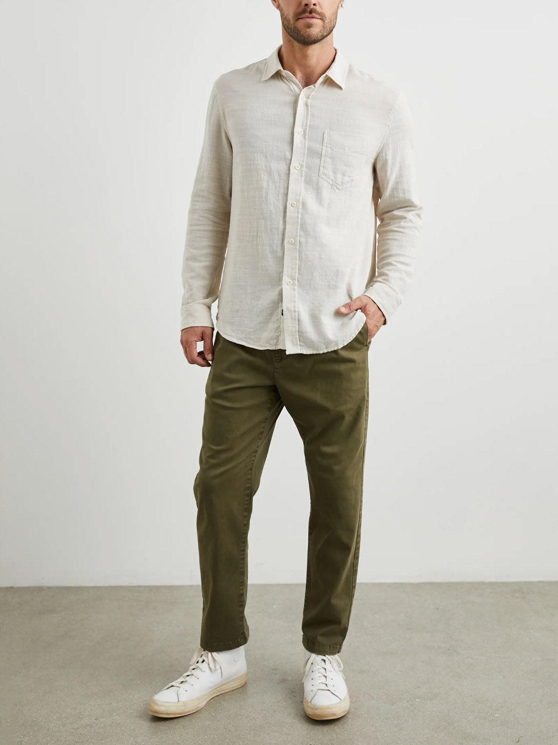 A person wearing an untucked Rails Wyatt Shirt by Rails in classic off-white, paired with olive green pants and white sneakers, stands against a plain light background. One hand is in the pocket, and their face is not visible.