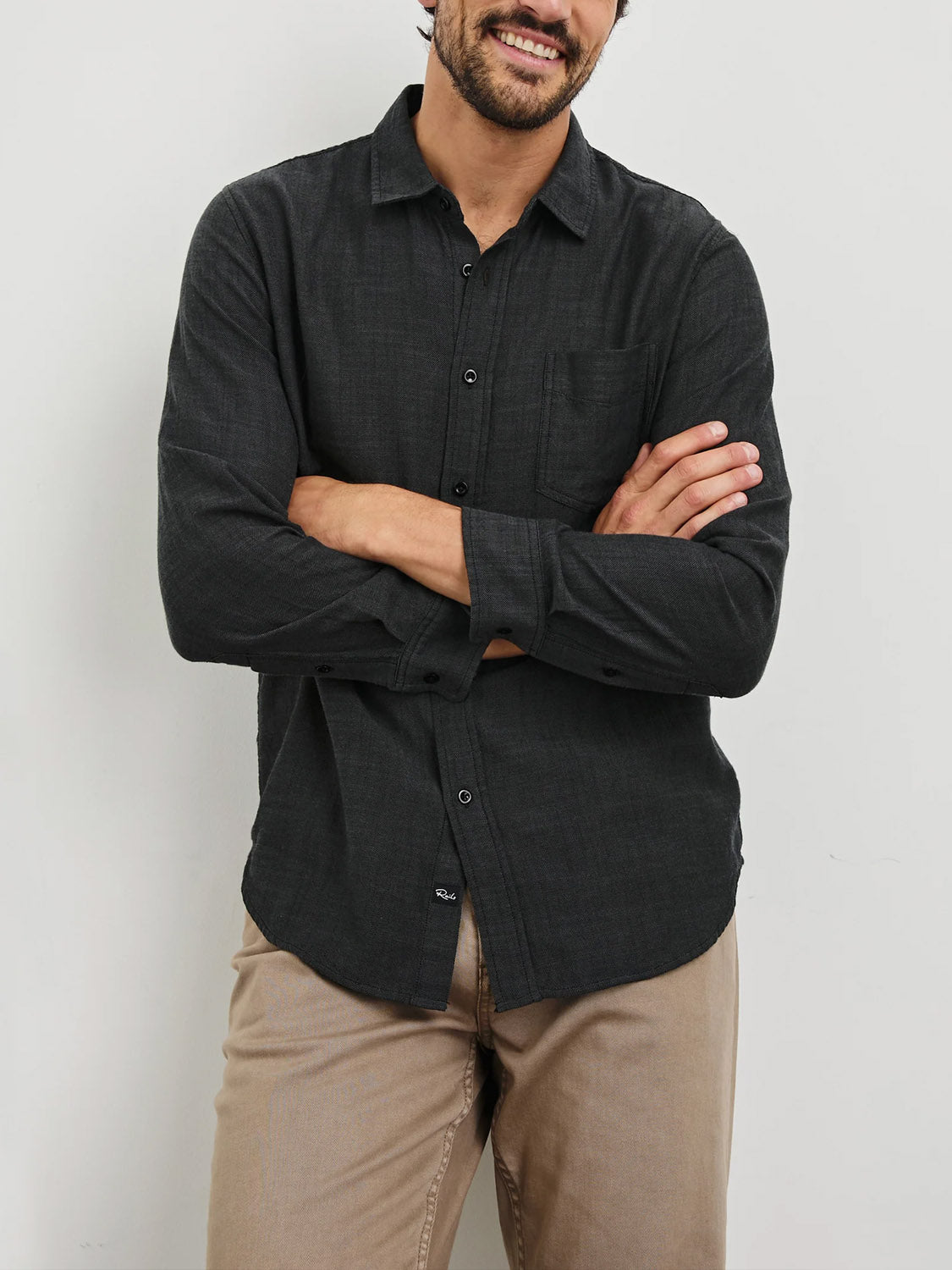 A man wearing a Rails Wyatt Shirt, a dark button-down made from cotton, and beige pants, stands with arms crossed, smiling against a plain light background.