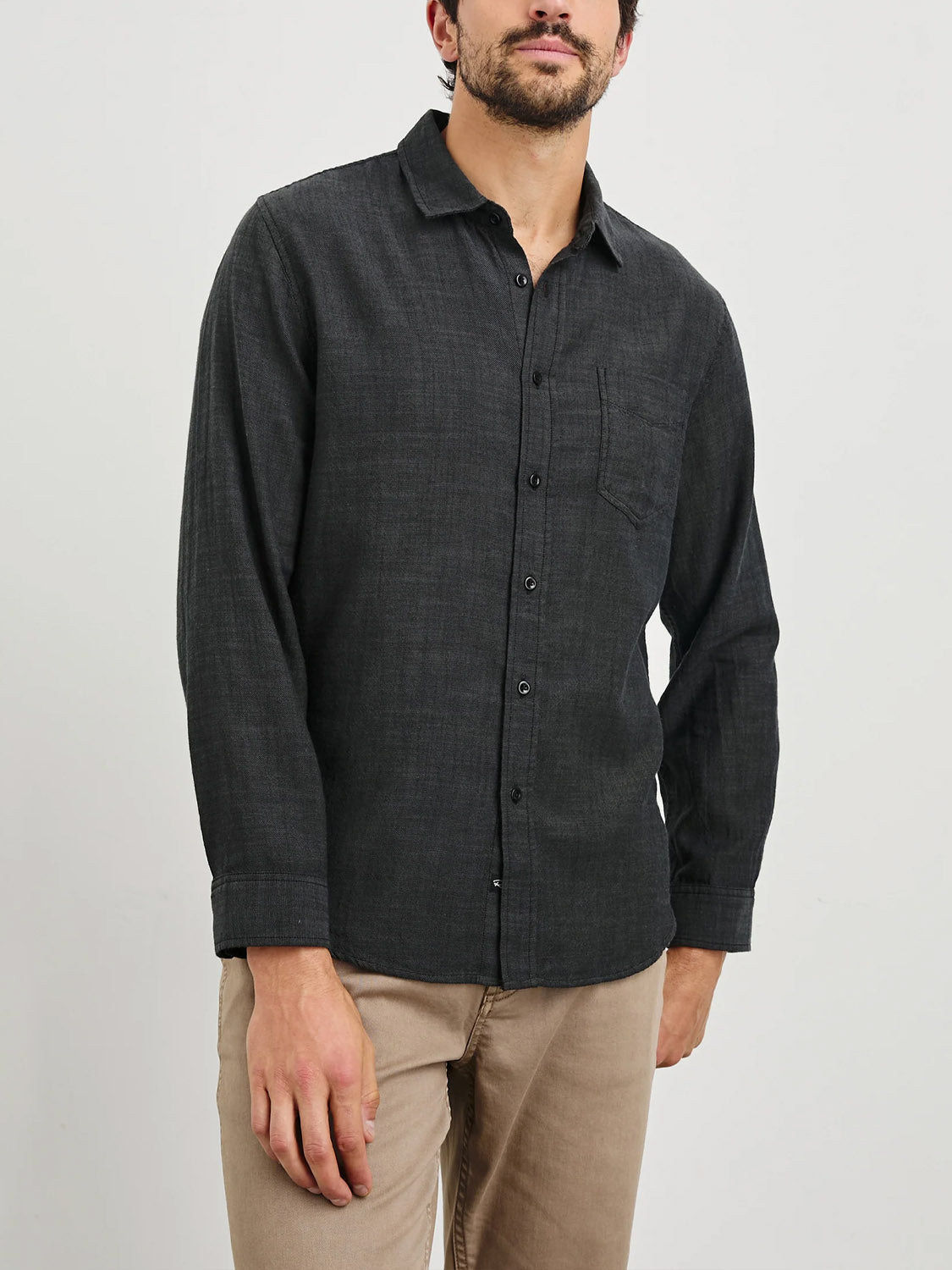 A person in a Rails Wyatt Shirt, a classic dark gray button-up made from textured cotton with a left chest pocket, stands against a white background, paired with light brown pants.