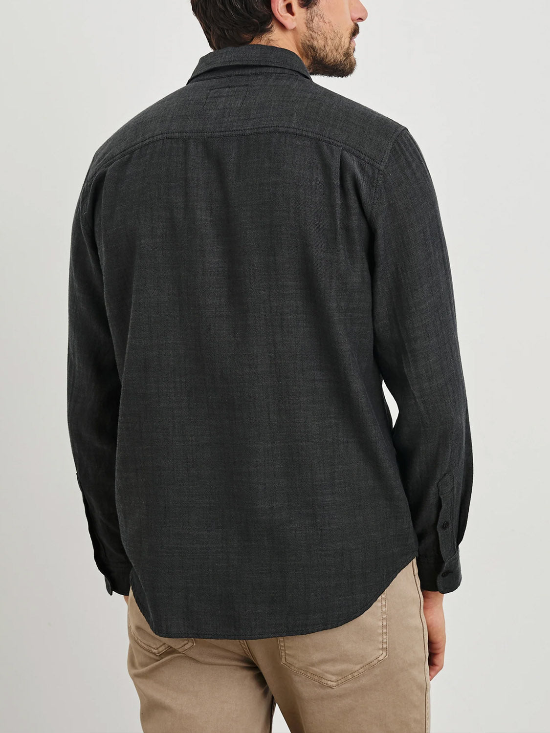 A man with short hair stands with his back to the camera, wearing a Rails Wyatt shirt in dark gray cotton paired with beige pants against a plain white background.