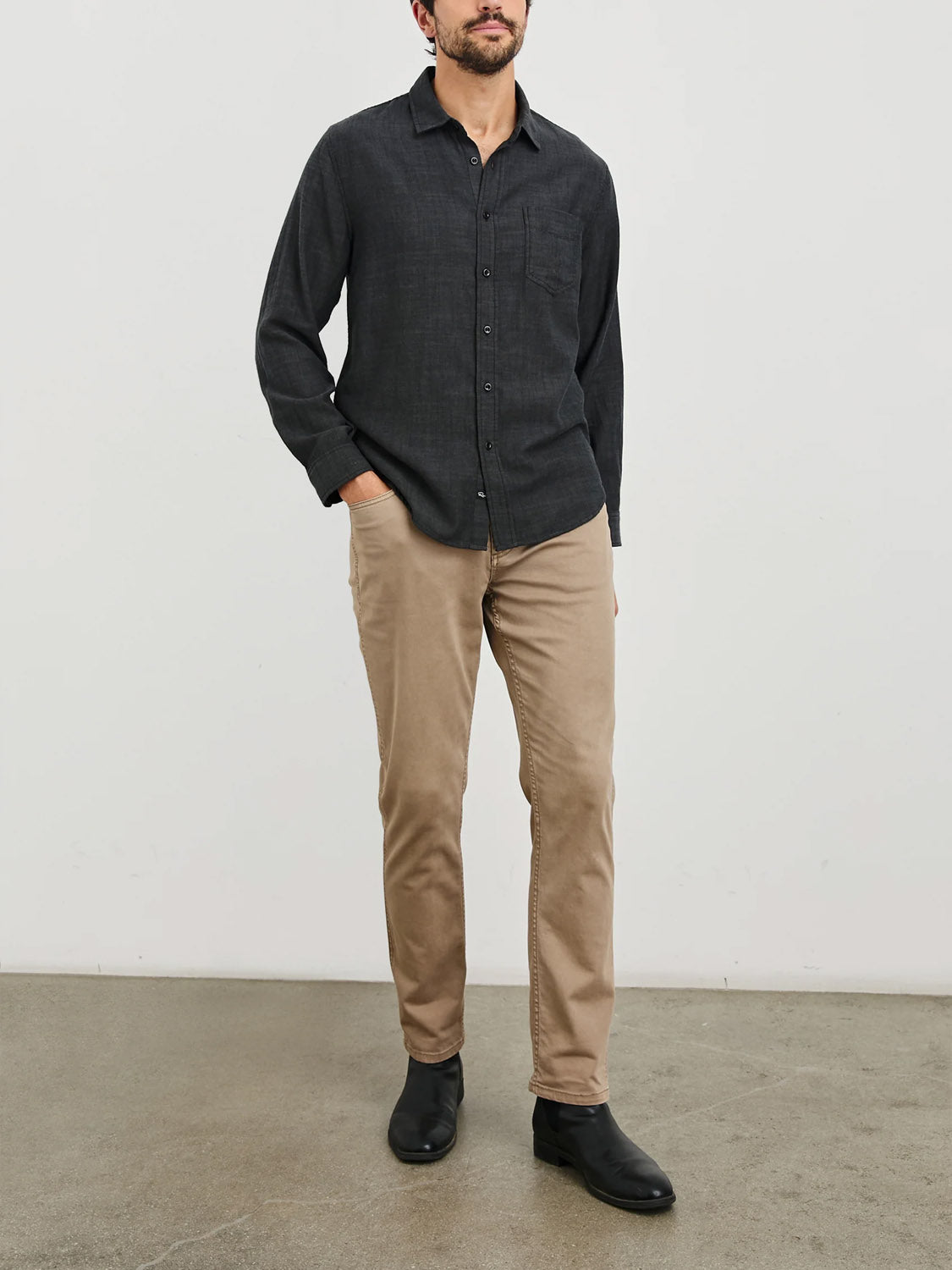 A person wearing a Rails Wyatt Shirt, beige pants, and black shoes stands against a plain light-colored wall on smooth, polished flooring. Their face is out of frame.