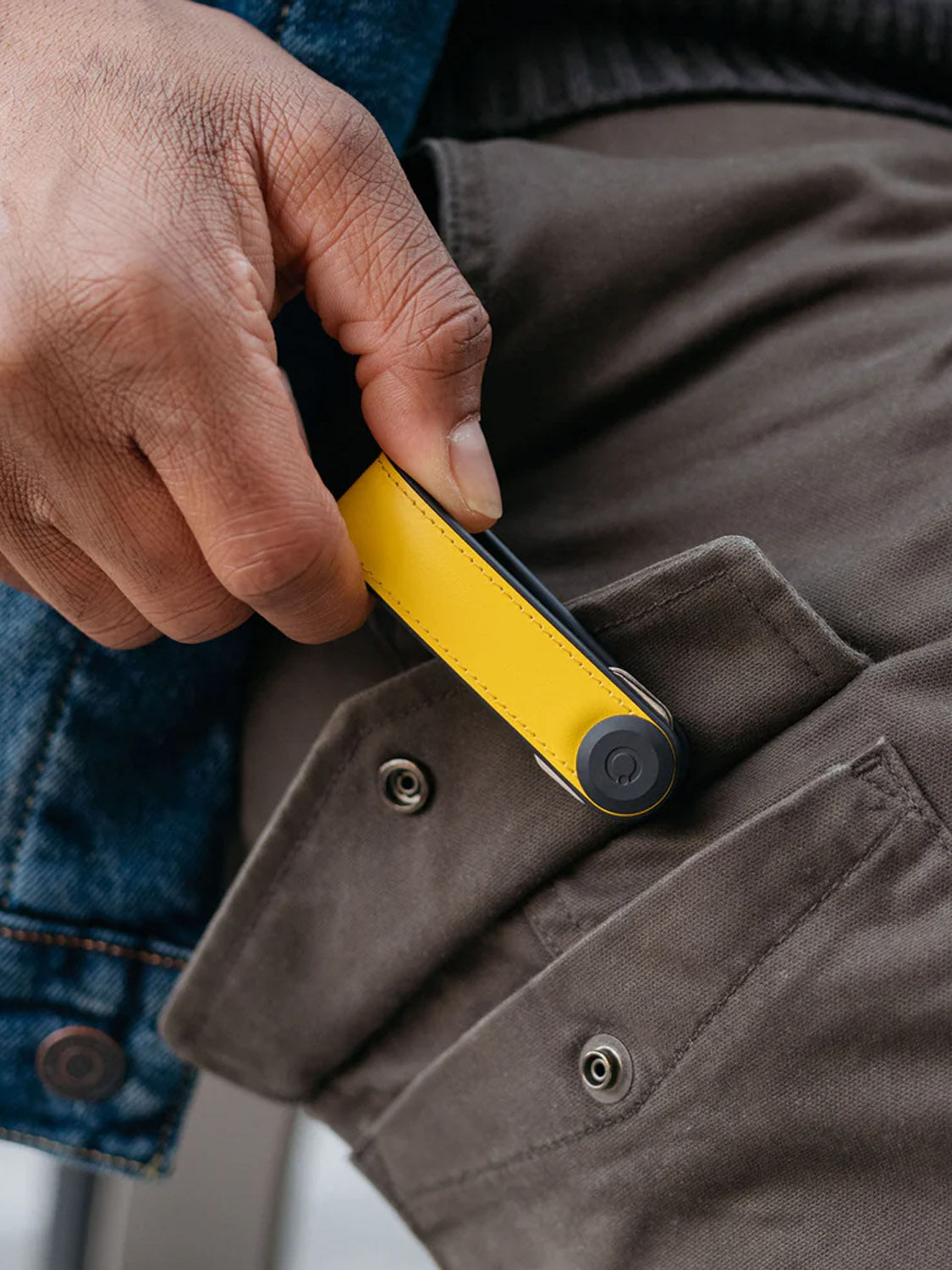 A hand holds the Orbitkeys Key Organizer Hybrid Leather in yellow, featuring a secure locking mechanism, and slides it into the front pocket of brown pants with visible buttons. The person is wearing a blue denim jacket.