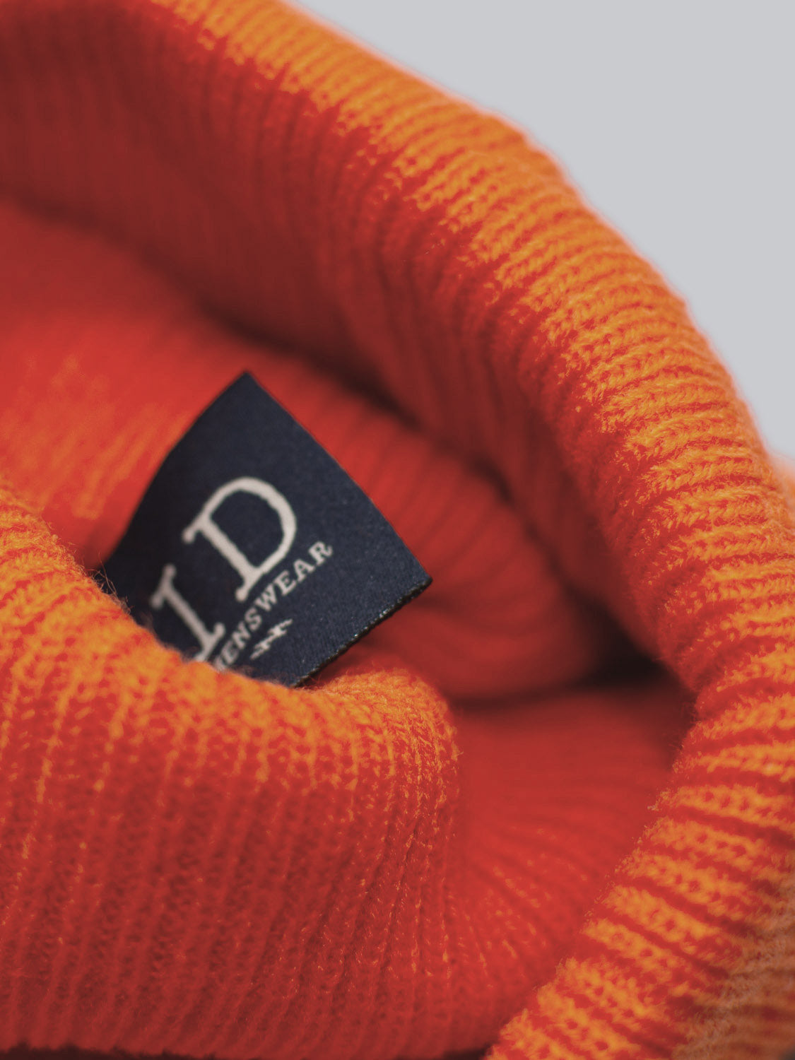 Close-up photo of the Yonker Ribbed Acrylic Beanie by ID Menswear in vivid orange, showcasing a blue label with partial branding. The soft texture and bright color epitomize this fisherman-style accessory.