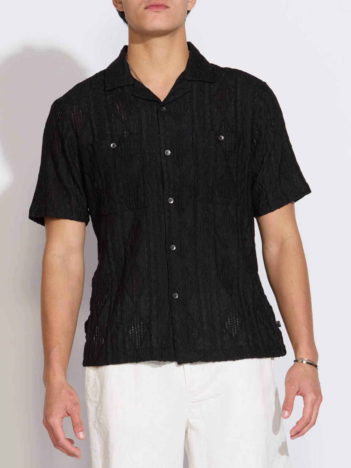 A model wears the Civil Society Zacatecas Jacquard Knit Resort Shirt, featuring a black jacquard knit, Cuban collar, and white buttons, paired with loose white pants. The shirt is shown from shoulders to hips against a plain background.
