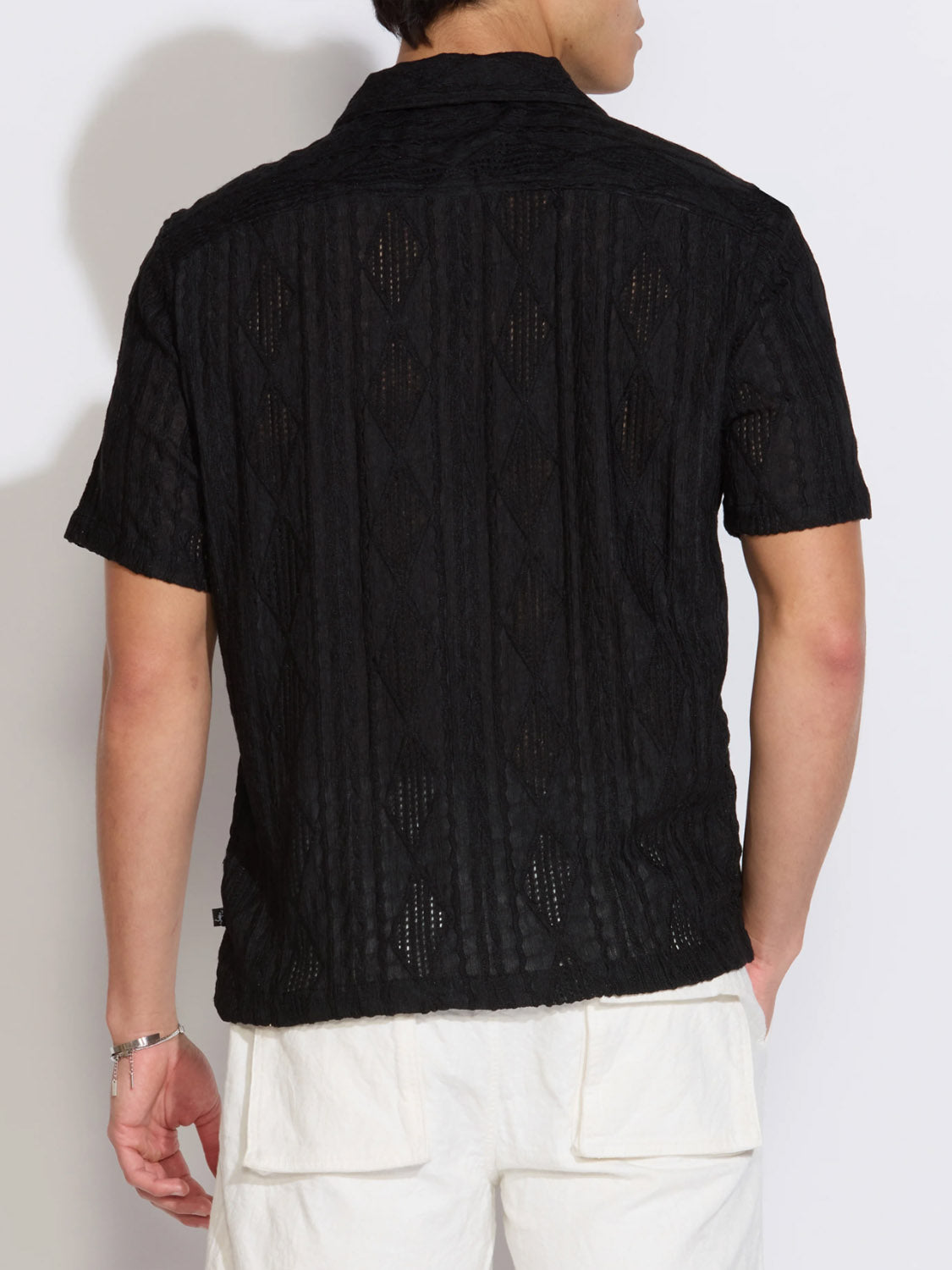 A person is seen from behind wearing the Civil Society Zacatecas Jacquard Knit Resort Shirt in black, featuring a diamond pattern and short sleeves, paired with white shorts against a plain light background.