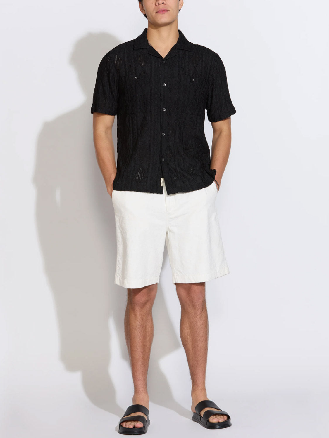 A person stands against a plain background wearing the Civil Society Zacatecas Jacquard Knit Resort Shirt, white knee-length shorts, and black slide sandals, with hands in pockets.