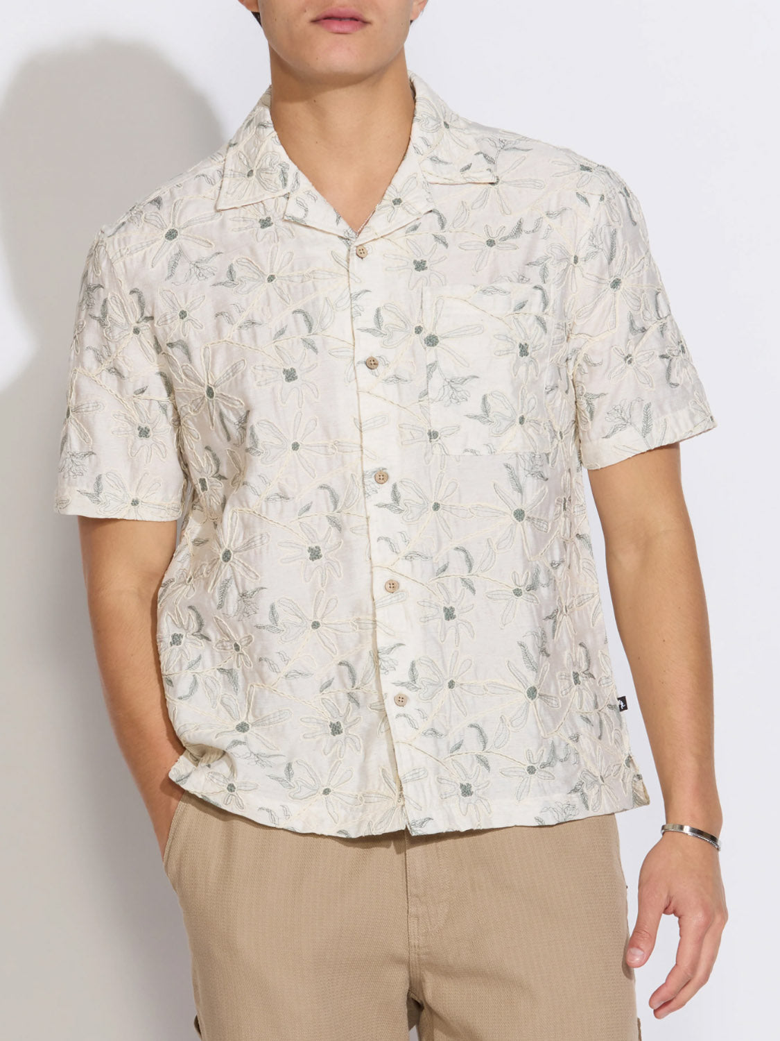 A person wears the Civil Society Zanzibar Embroidered Appliqué Resort Shirt—a light-colored, short-sleeve Cuban collar shirt with floral embroidery—paired with beige pants, standing with one hand in their pocket against a plain background.